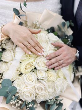 Close-up of bride and groom's hands with wedding rings resting on a white rose bouquet.