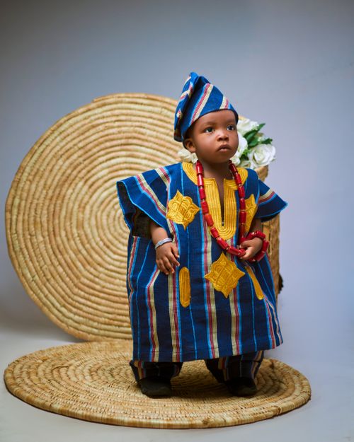 Free A young child dressed in vibrant traditional African clothing, standing against a woven backdrop. Stock Photo