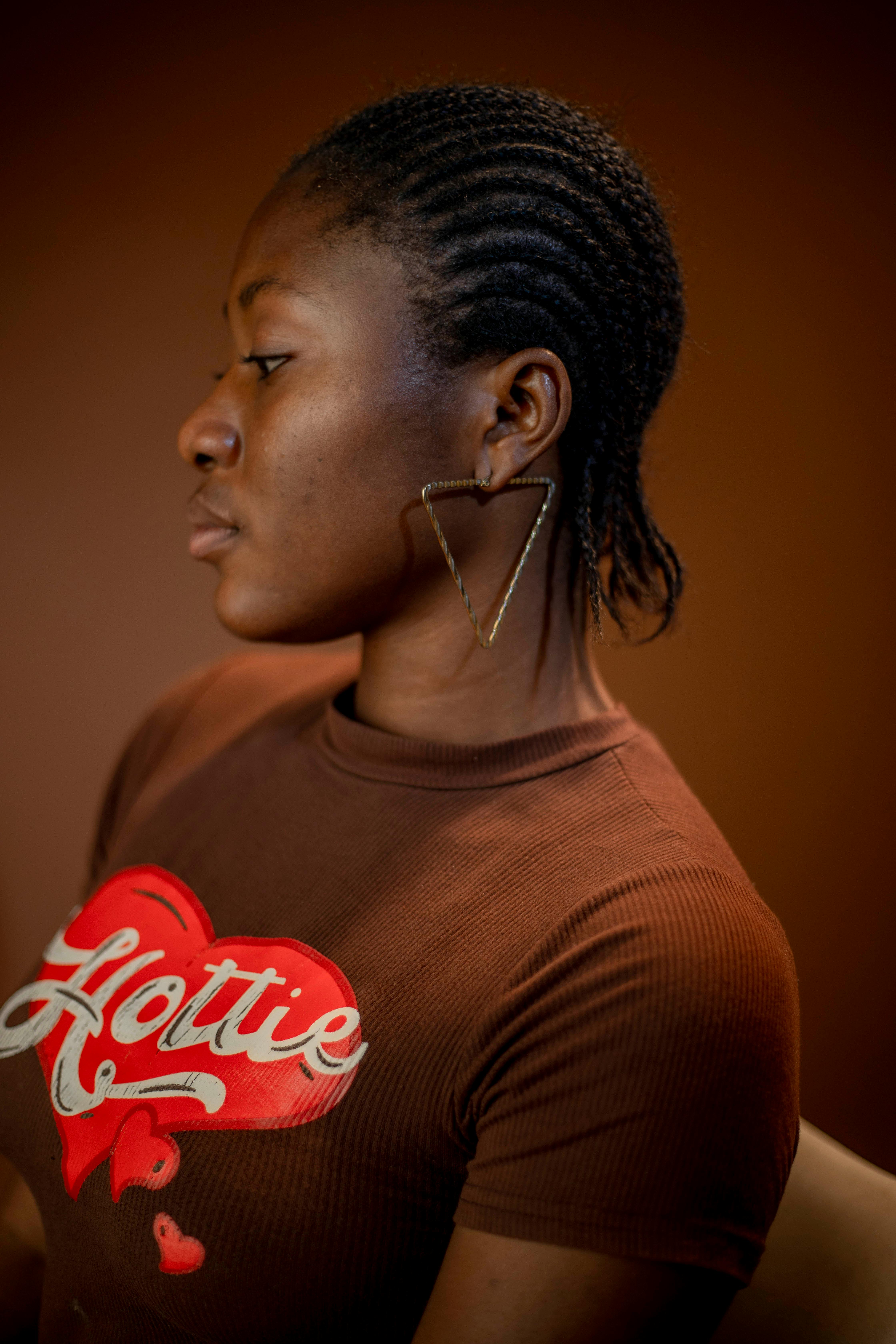 Side profile of a woman with braided hair wearing a brown T-shirt and earrings in a studio setting.