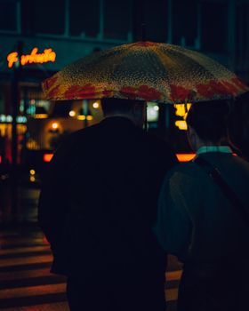 Couple walking under floral umbrella at night on a rainy street with neon lights.