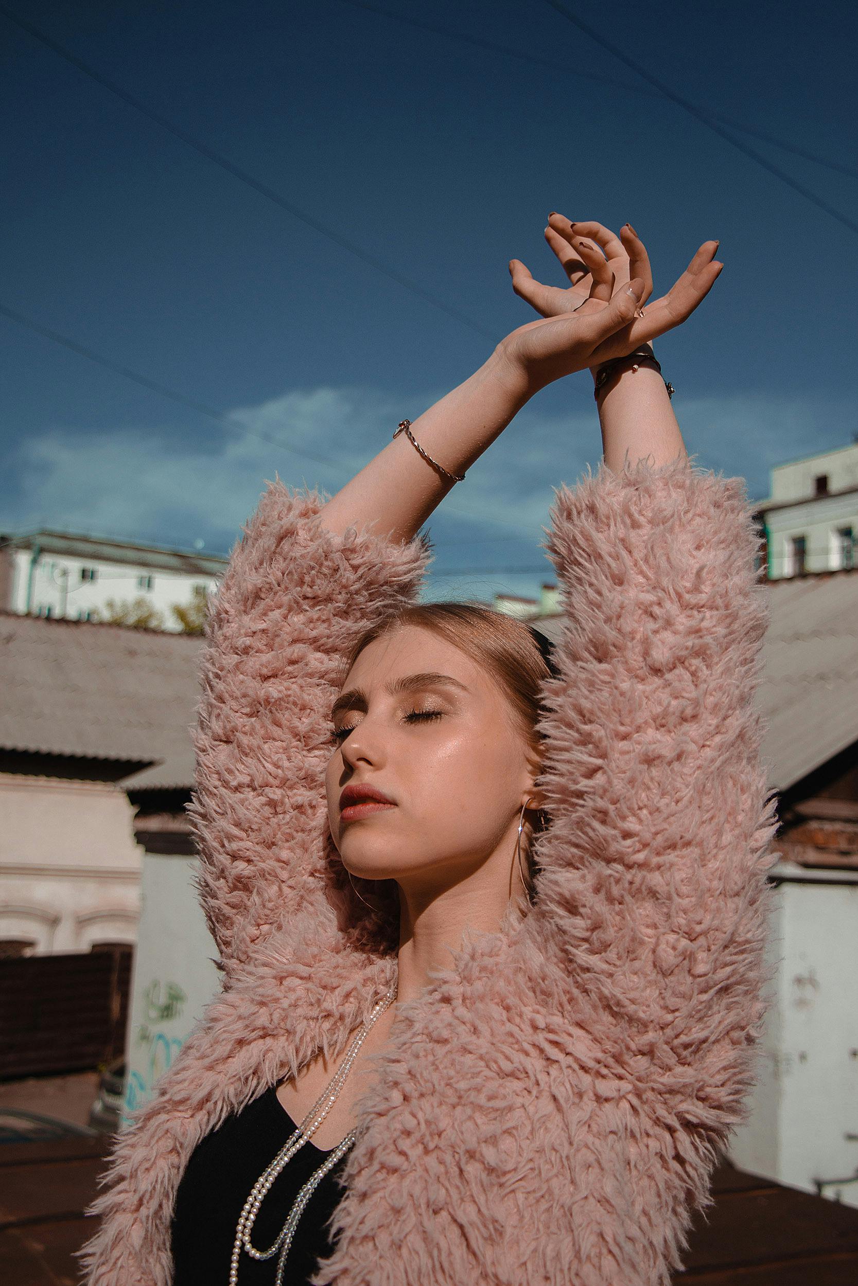 Fashionable young woman in a fluffy pink coat posing confidently outdoors with arms raised.