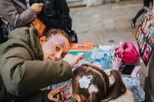 A face painter artistically decorates a child's face outdoors at a vibrant street festival.