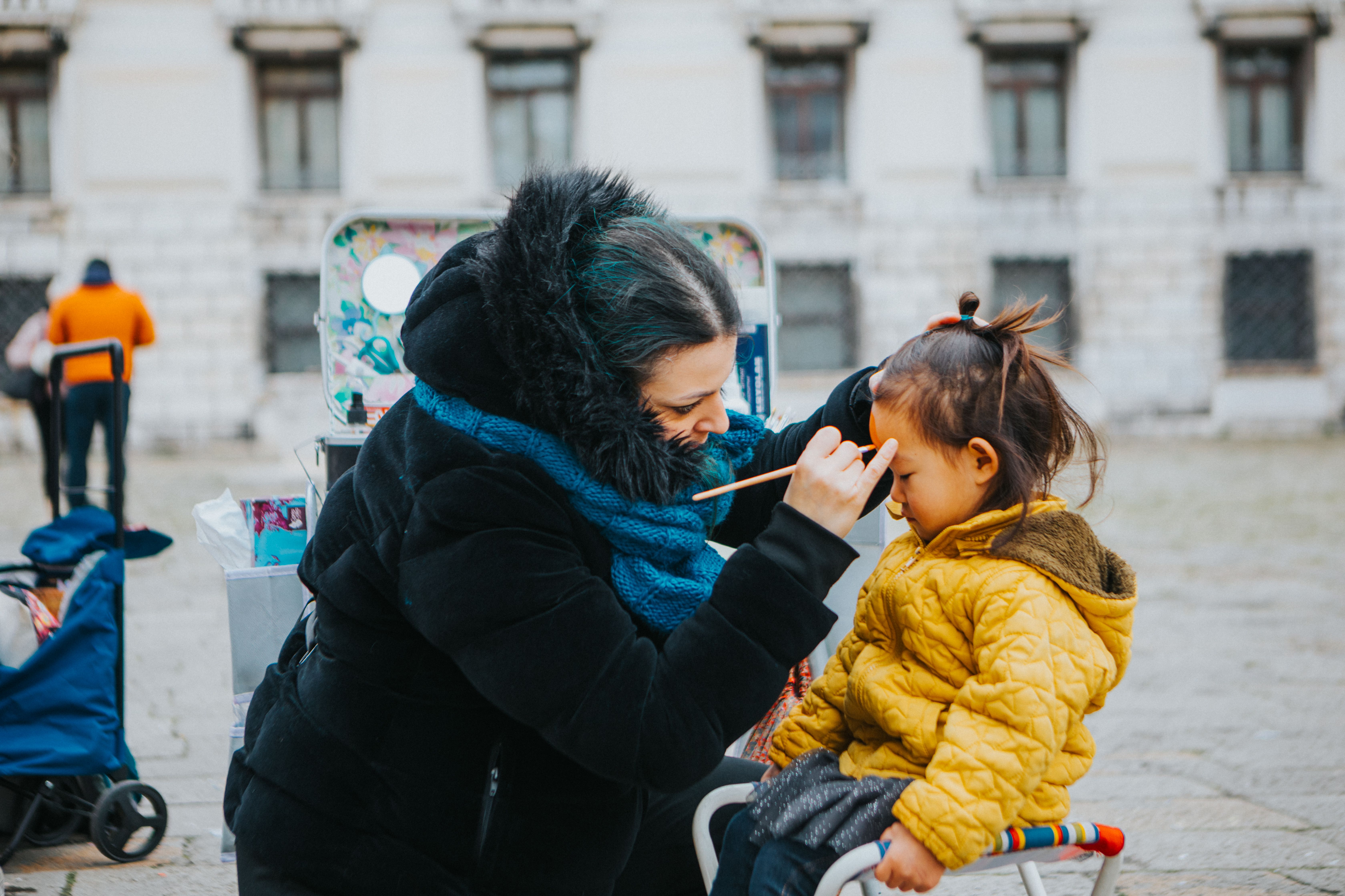 A face painter applying makeup to a young child outdoors in a public space, capturing a warm interaction.