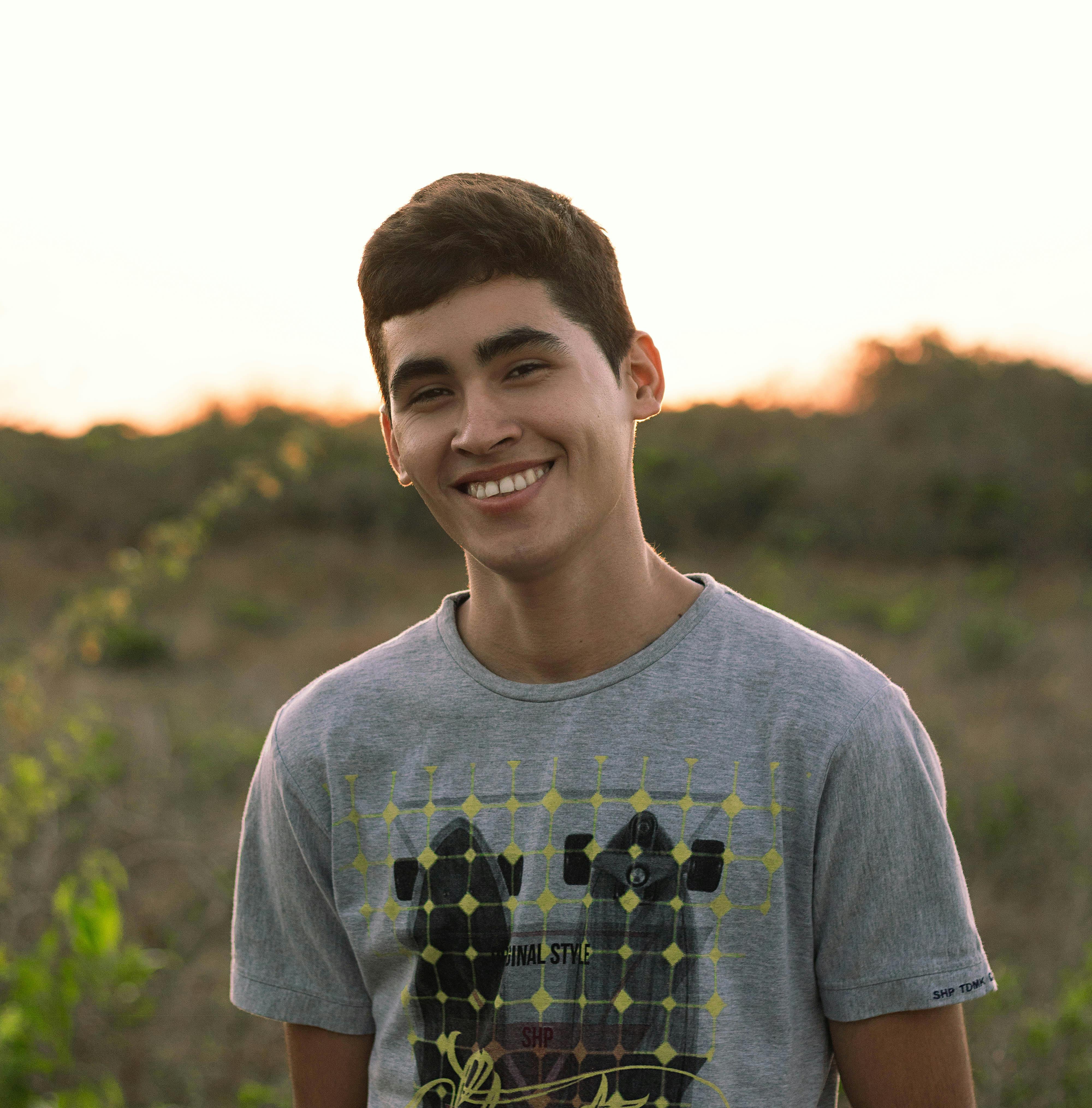 Positive young male in casual outfit smiling brightly while standing in grassy meadow in evening