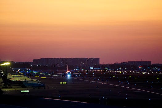 Airplane preparing for takeoff on an illuminated runway, against a sunset cityscape backdrop.