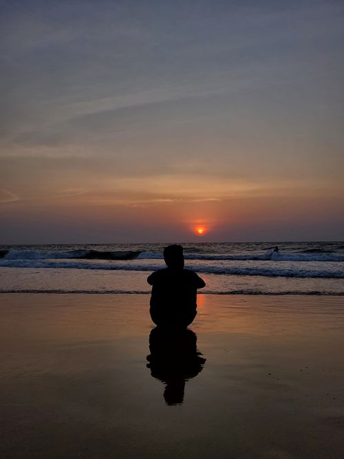 Free Silhouette of an individual seated on the beach facing a vibrant sunset over the ocean. Stock Photo
