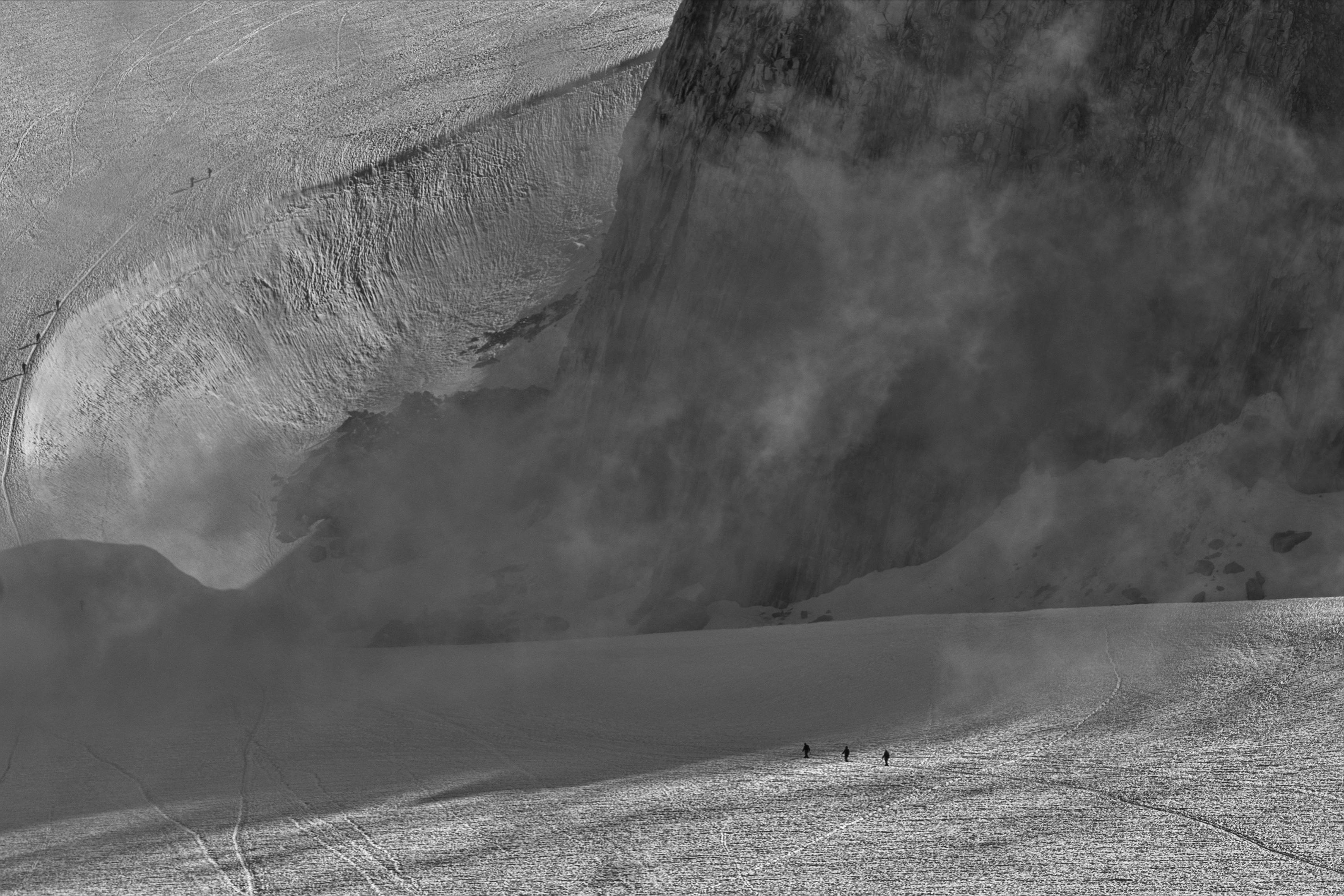 Remote hikers traverse a snowy mountain under a moody sky.