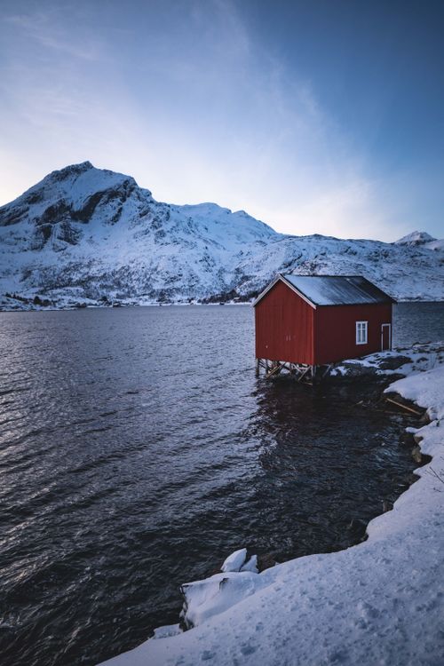 Free A picturesque red cabin by a snowy lake with majestic mountains in winter. Stock Photo