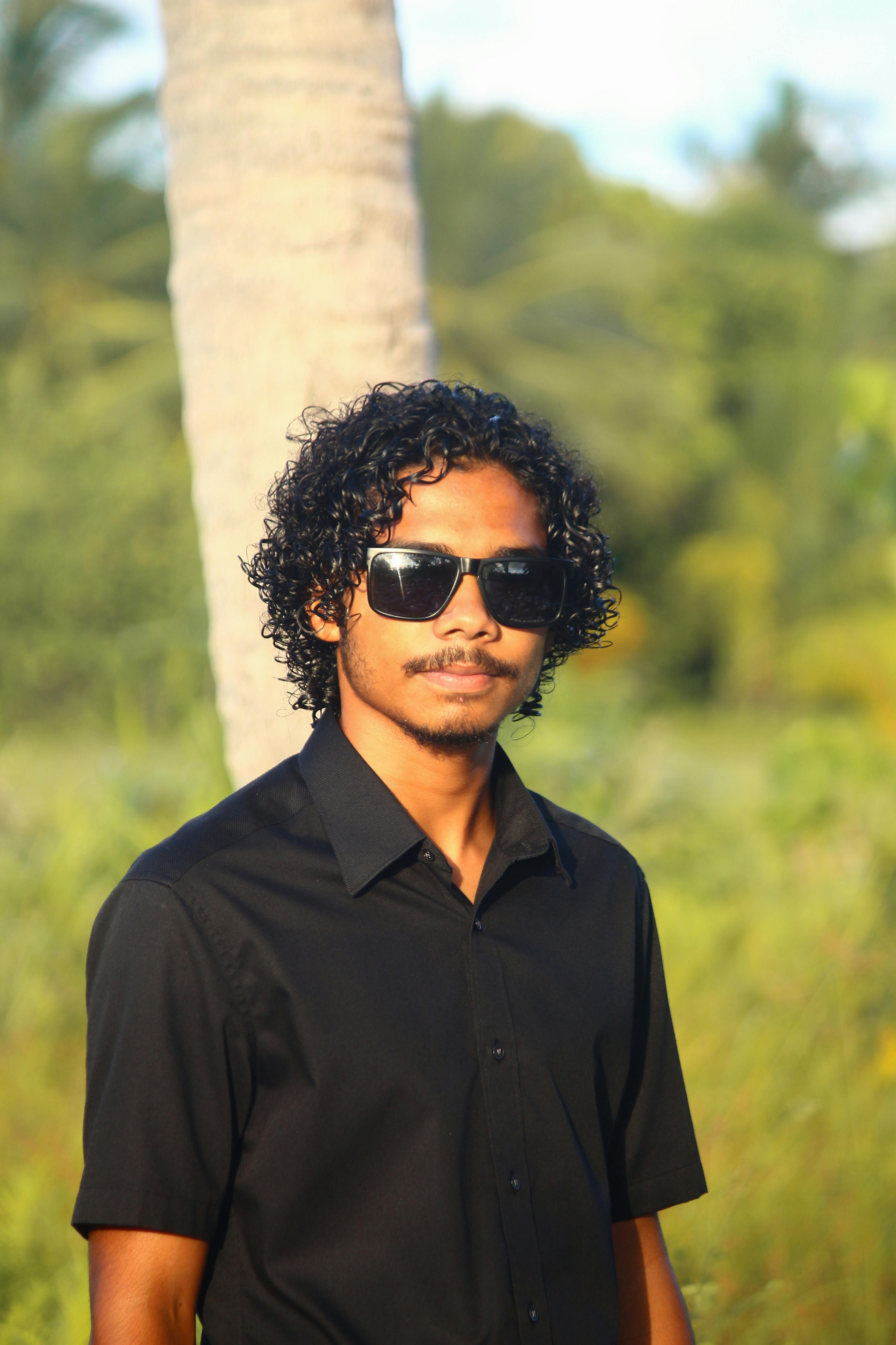 Portrait of a young man with curly hair and sunglasses in a natural setting with palm trees.