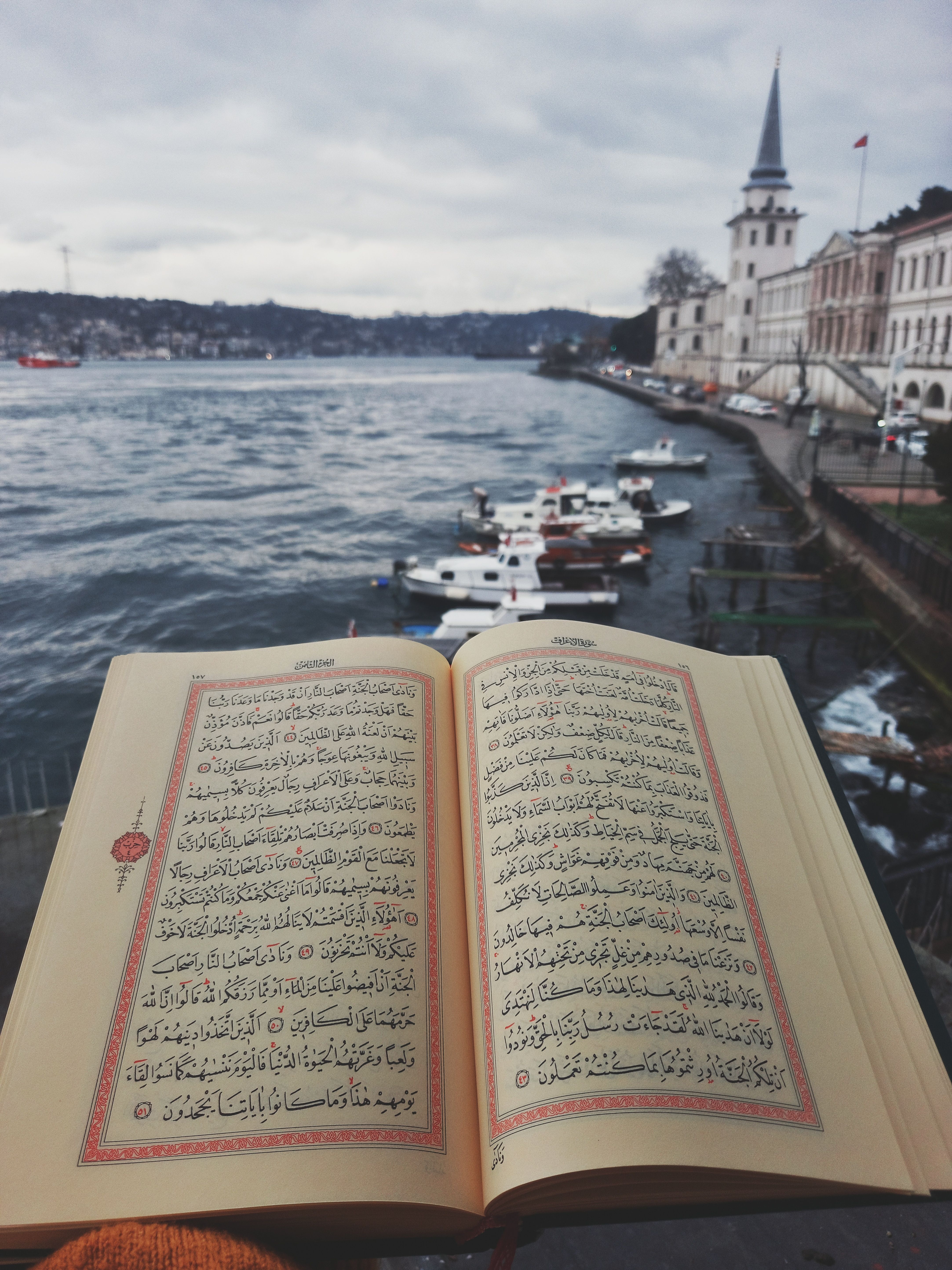 Quran open with Arabic text in front of a scenic waterfront mosque view.