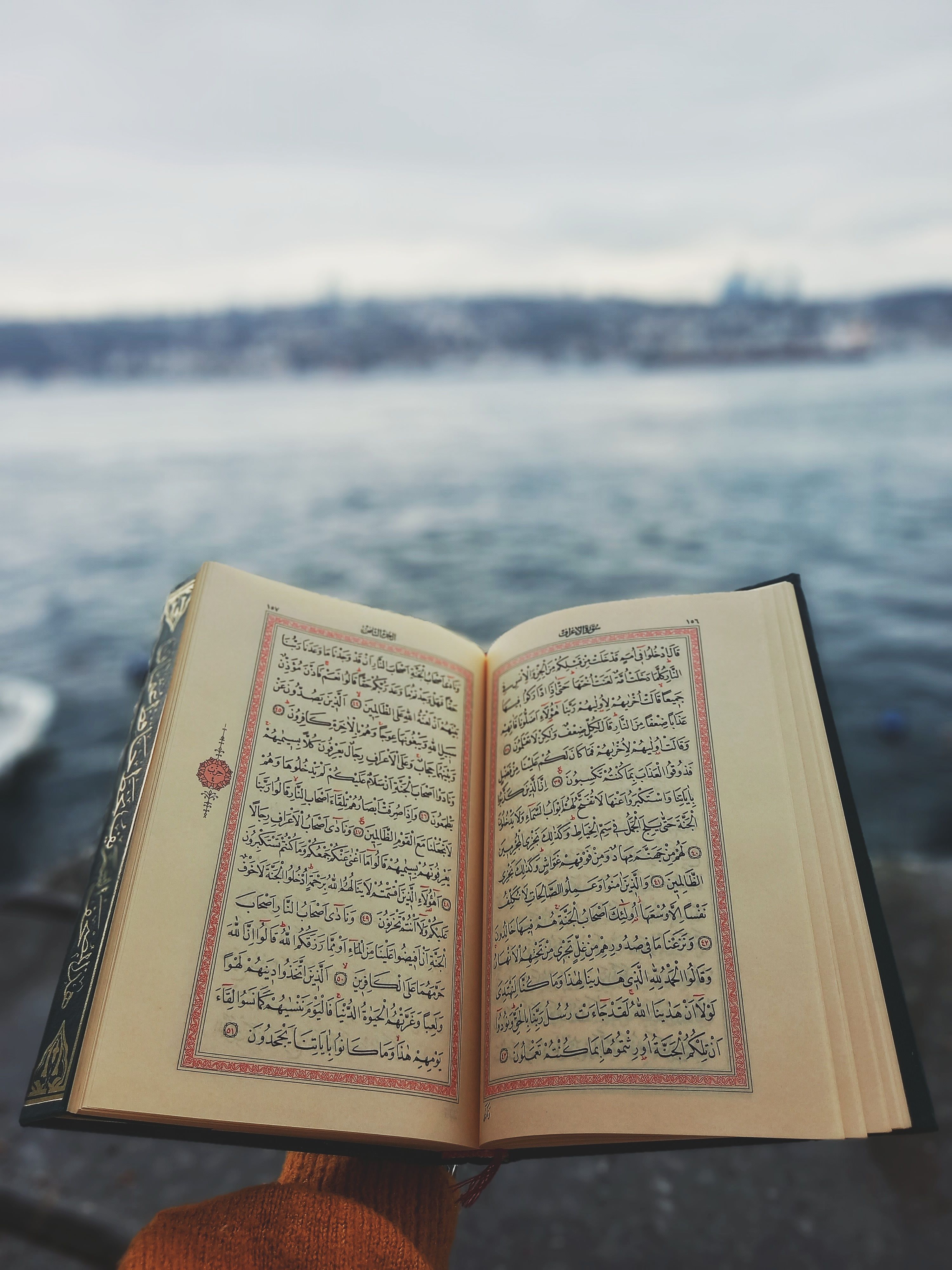 Open Quran being held on a riverbank with city skyline in the background.