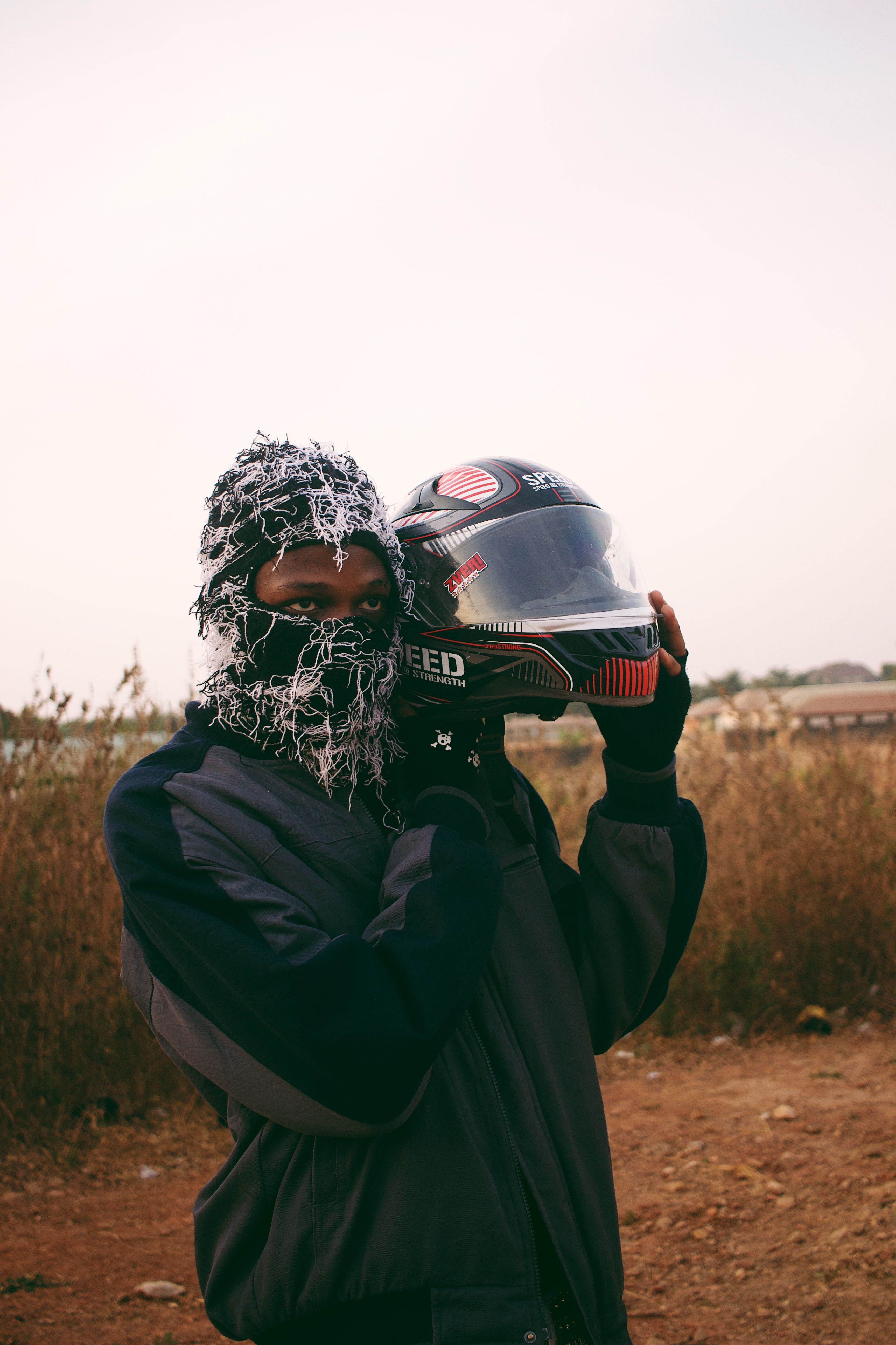 A person wearing winter gear holding a motorcycle helmet outdoors in Nigeria.