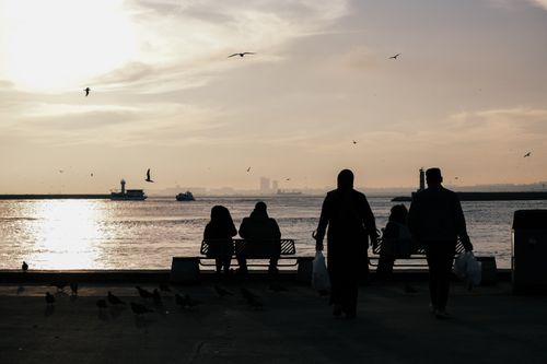 Free A peaceful sunset over water with silhouettes of people sitting and walking along the waterfront. Stock Photo