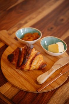 Freshly baked croissant on a wooden board with butter and apricot jam.