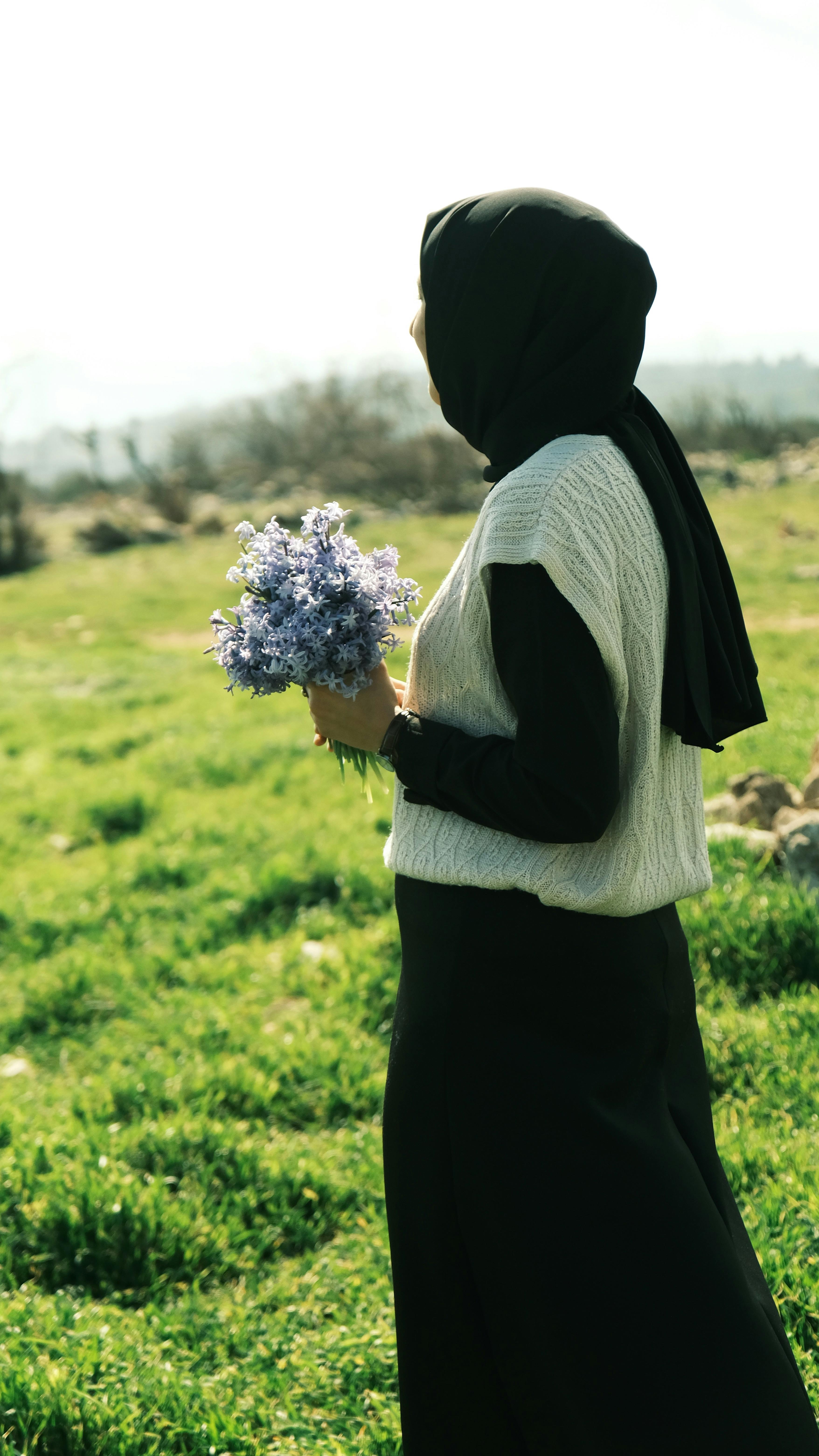 A woman in hijab holds a lavender bouquet in a sunny, grassy meadow.