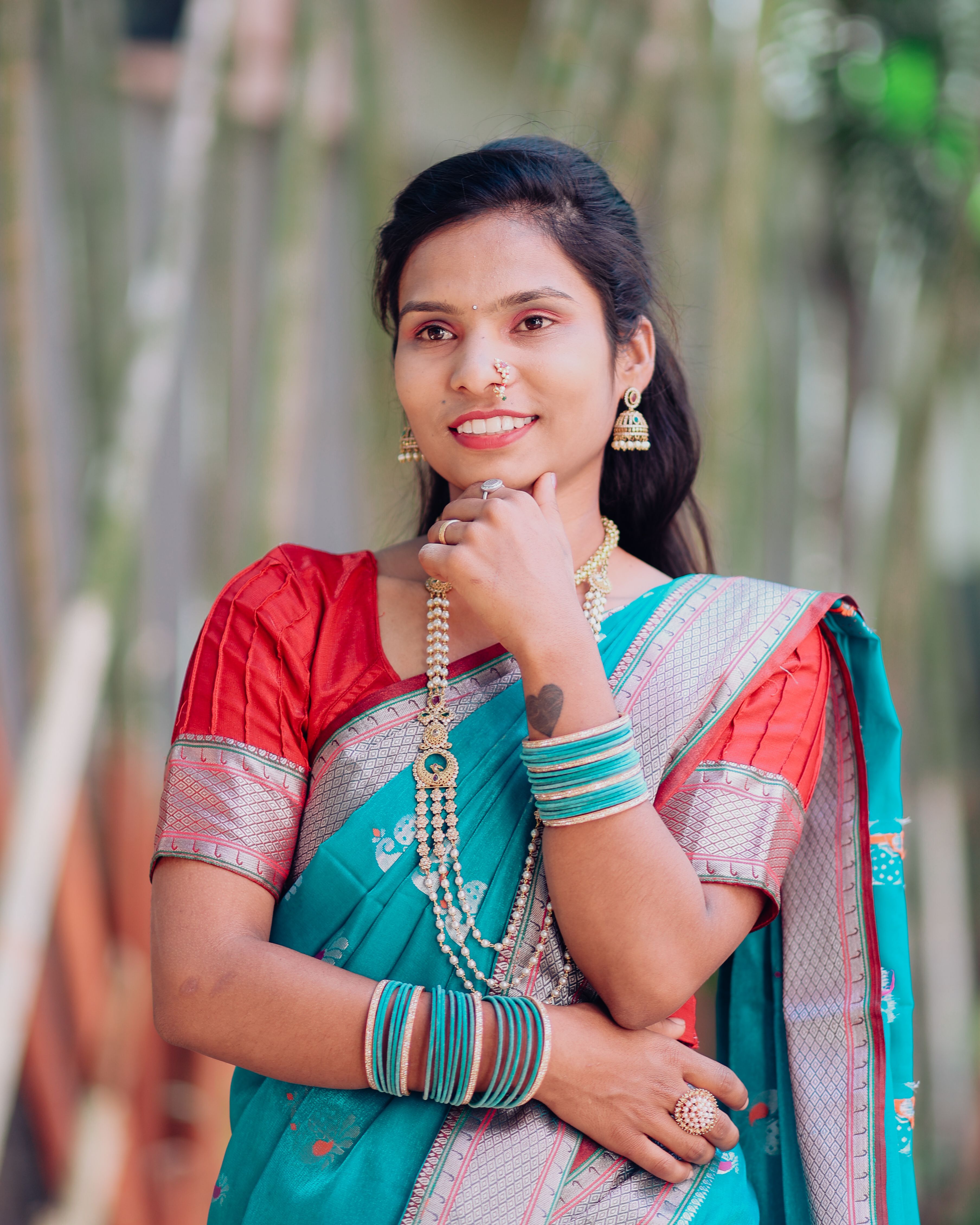 A woman in a colorful saree and jewelry smiling outdoors with a serene background.