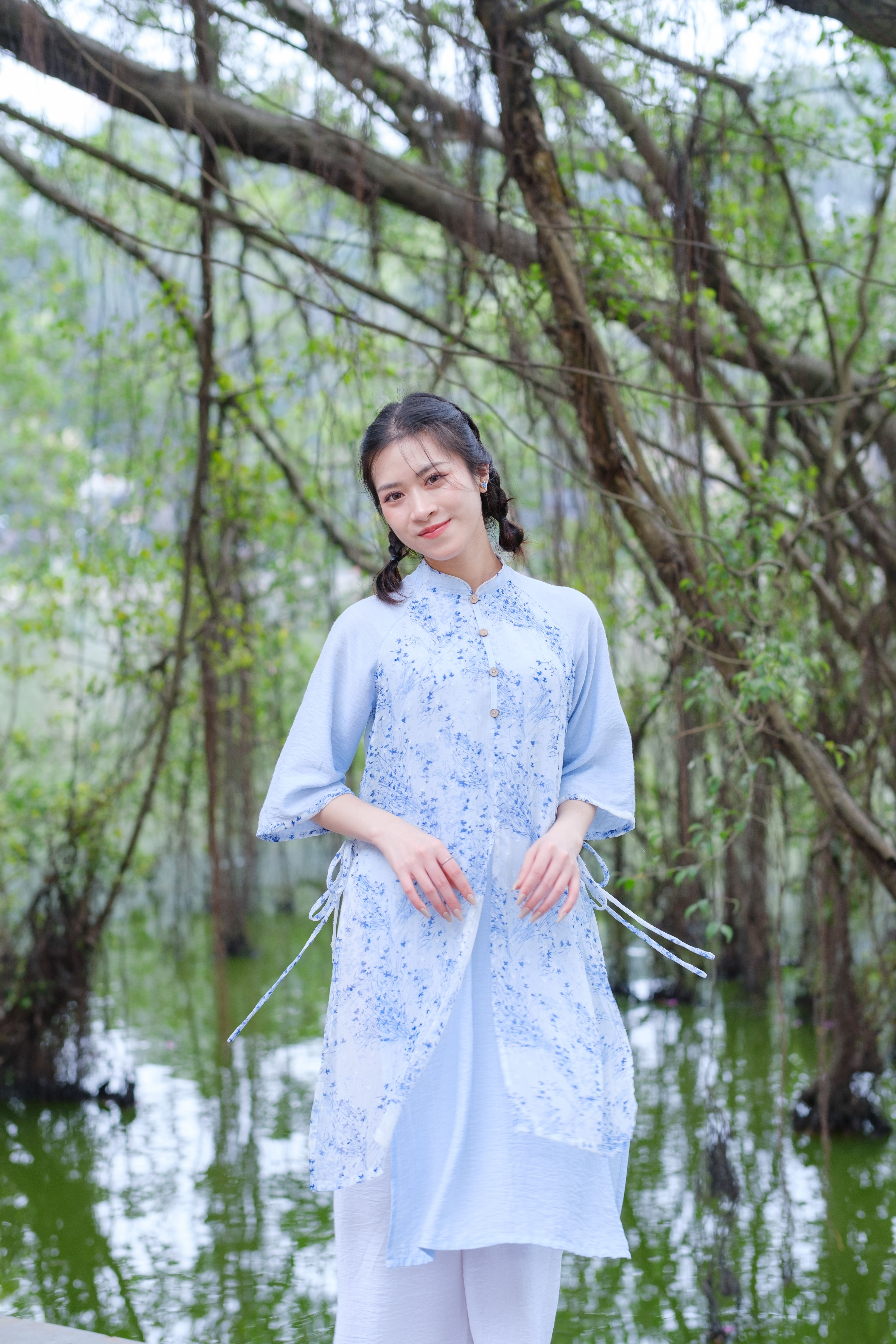 A woman stands in a traditional dress by a serene lagoon, surrounded by lush greenery.