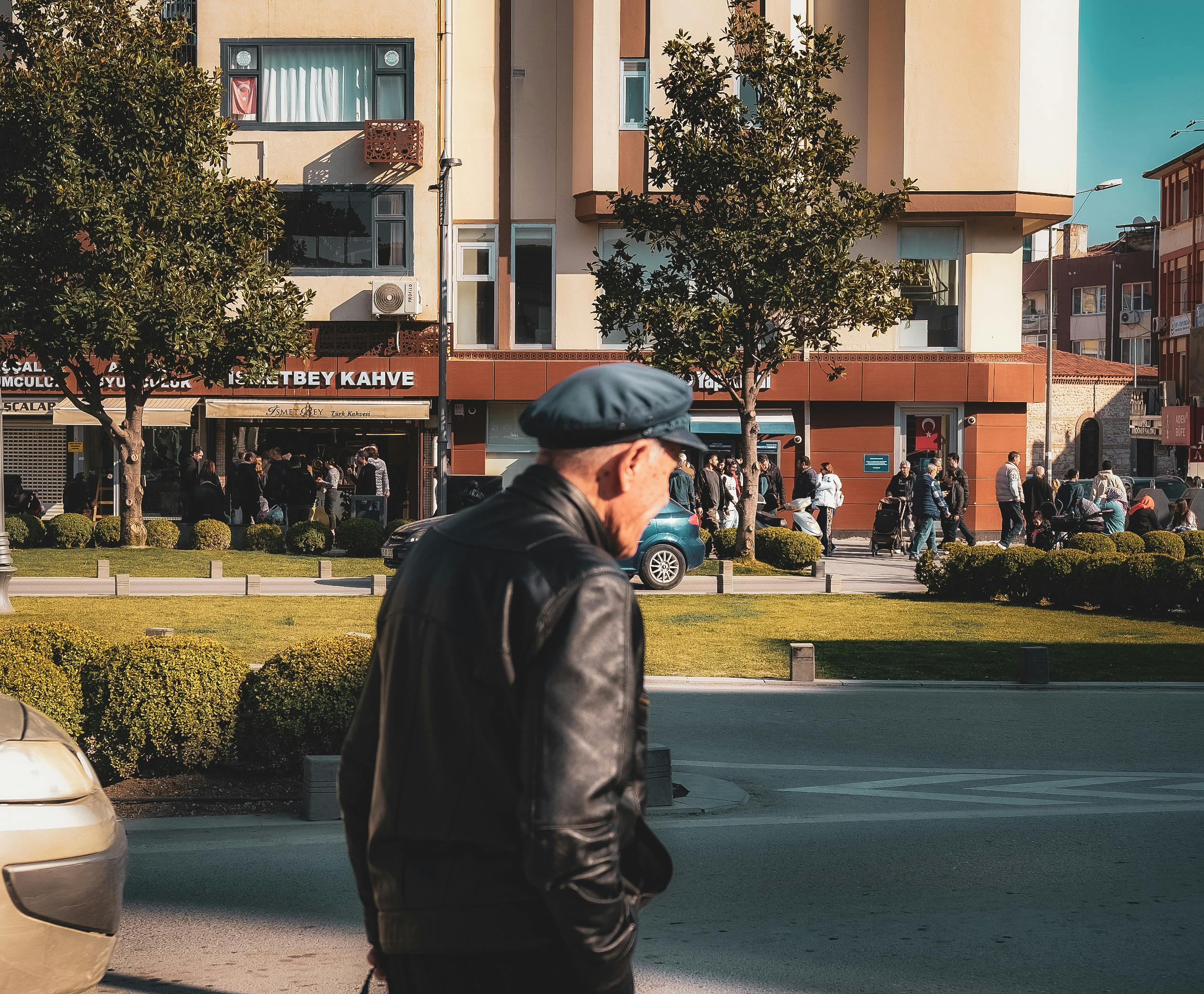 Elderly man in a leather jacket walks on a bustling city street on a sunny day.