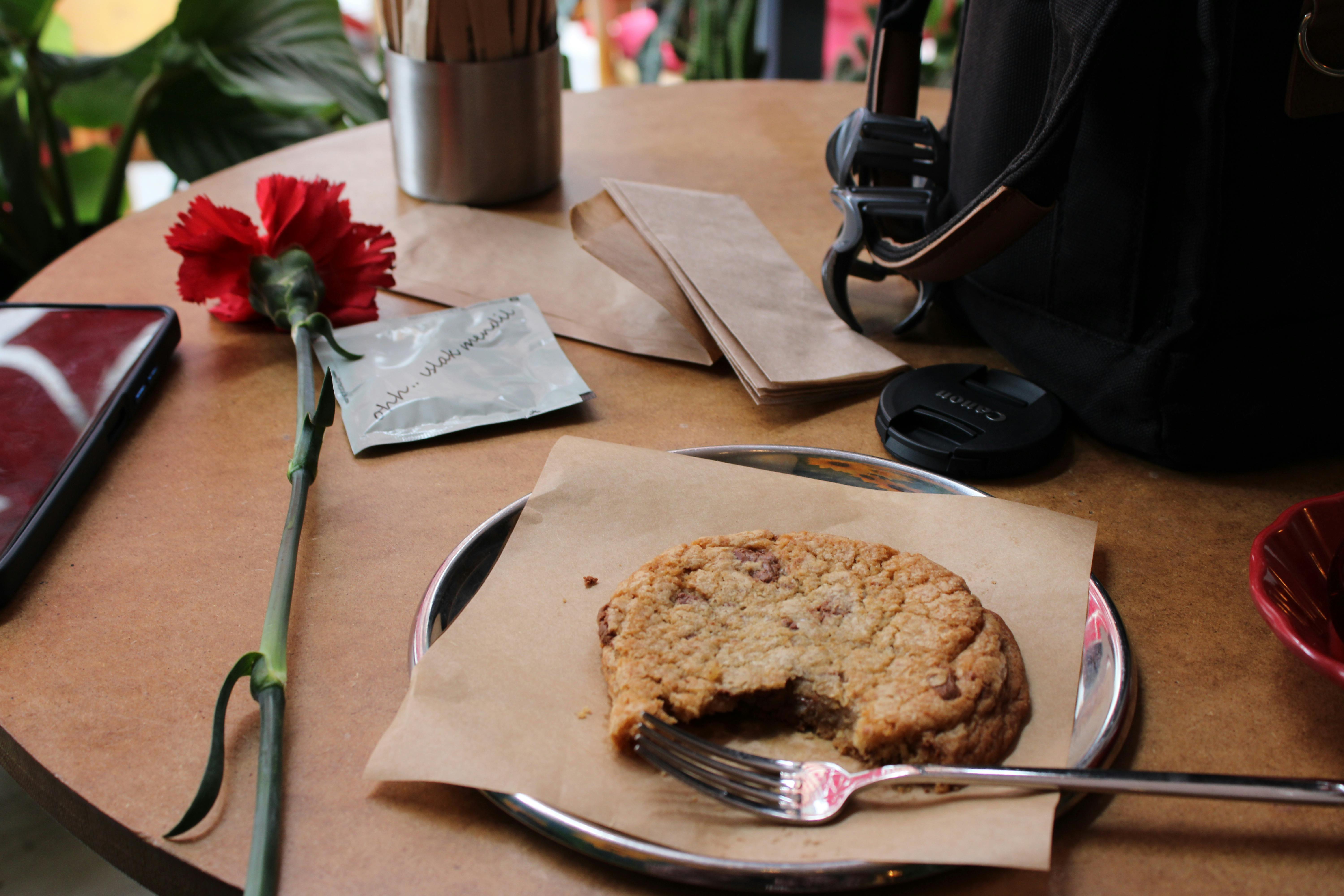 A cozy cafe setup with a chocolate chip cookie and a red carnation on a table.