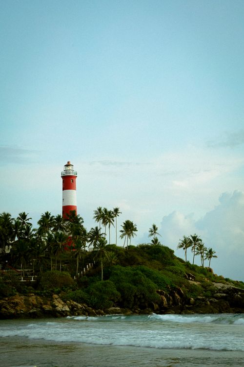 Free A scenic view of a red and white lighthouse surrounded by tropical palm trees by the ocean. Stock Photo