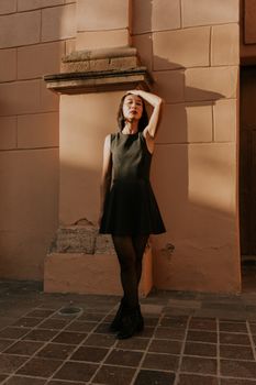 Elegant woman in black dress poses against sunlit wall, creating striking shadows.