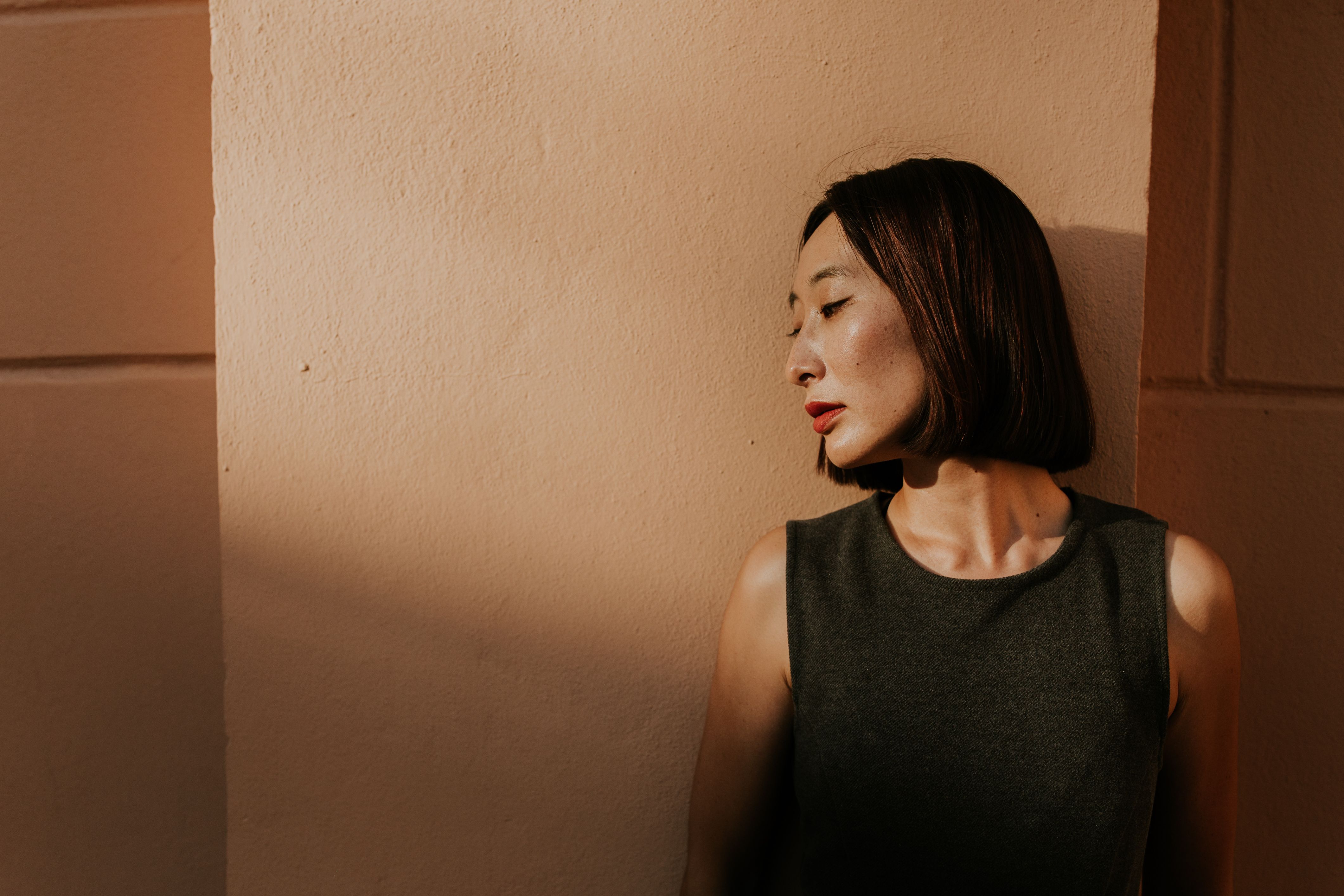 A woman leans against a sunlit wall creating abstract shadows in a serene setting.