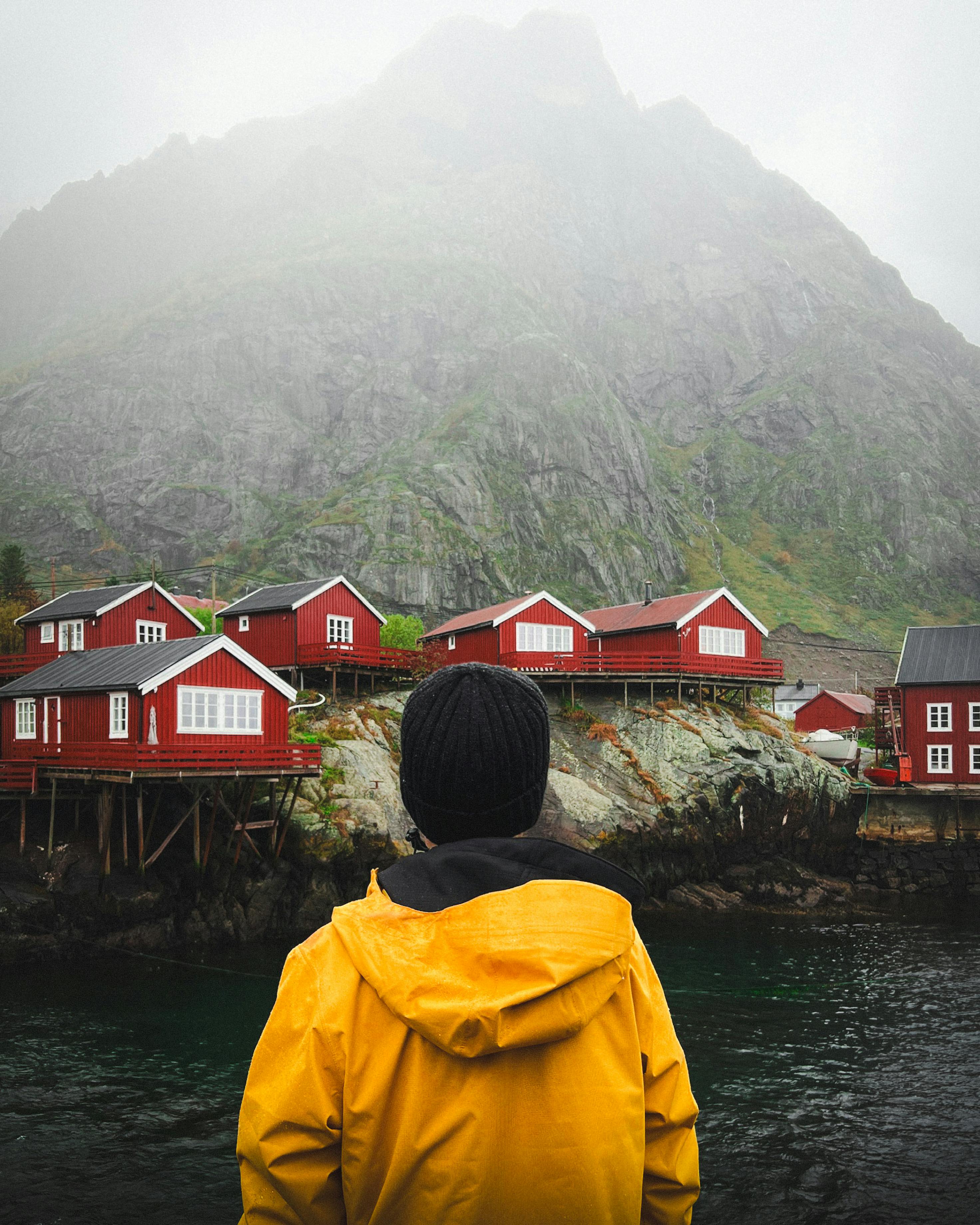 Person in yellow jacket overlooking red cabins in foggy Lofoten, Norway.
