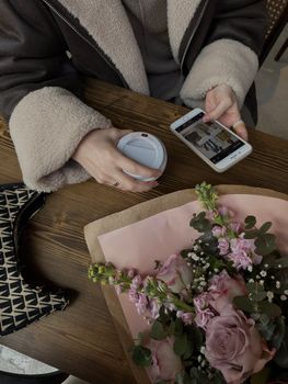 A cozy indoor scene with a woman holding coffee and browsing a phone beside pink flowers.