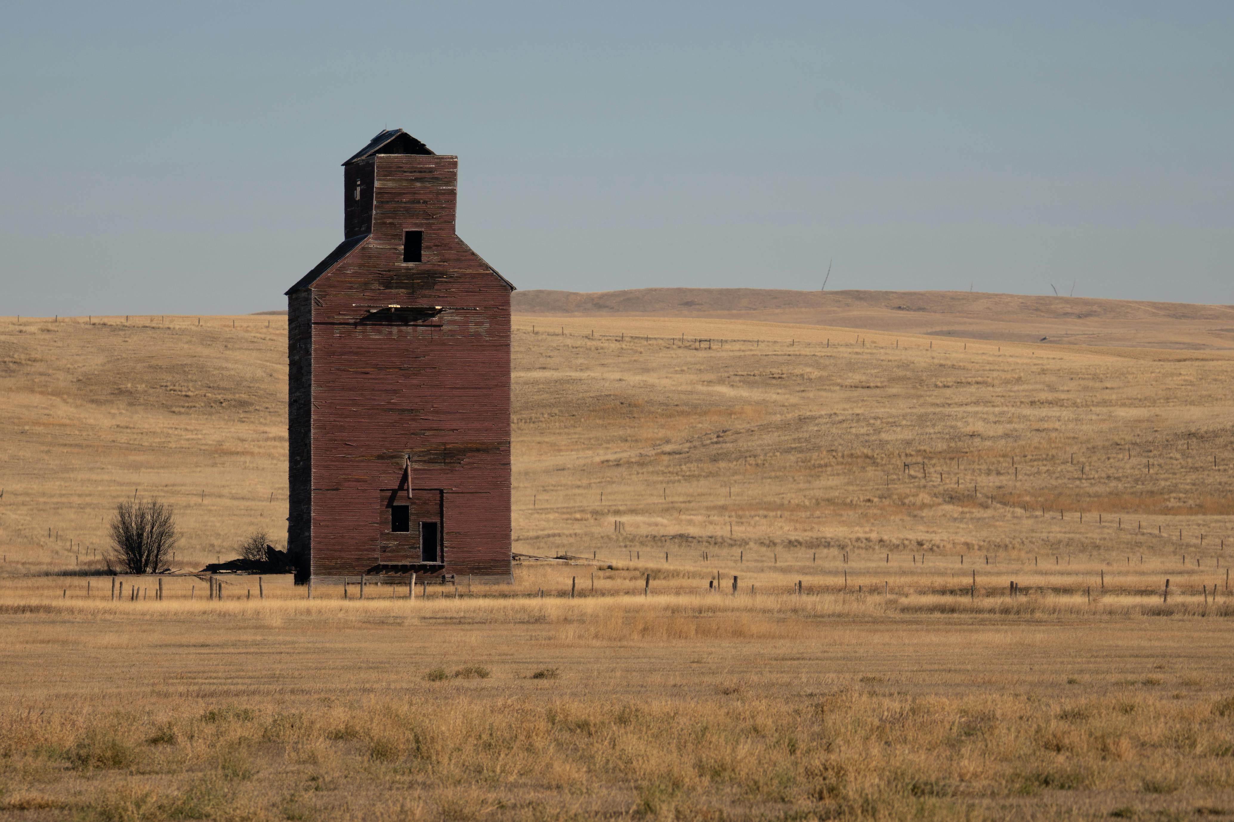 A weathered red barn stands isolated on a vast, dry grassland under a clear sky.