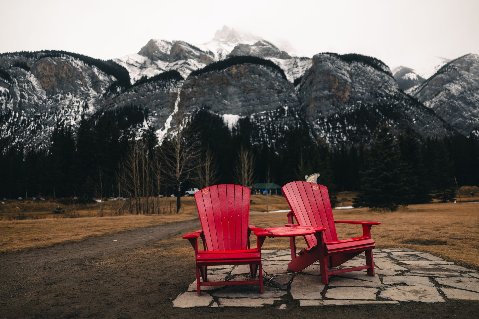 Two red Adirondack chairs on a stone path with a view of snow-capped mountains in Banff, Alberta.