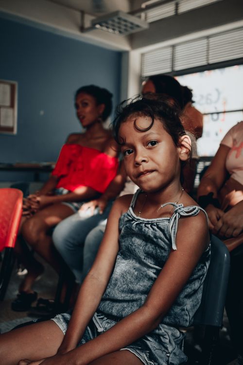 Free Calm preteen ethnic dark skinned girl in casual gray dress looking at camera while sitting on chair in public place Stock Photo