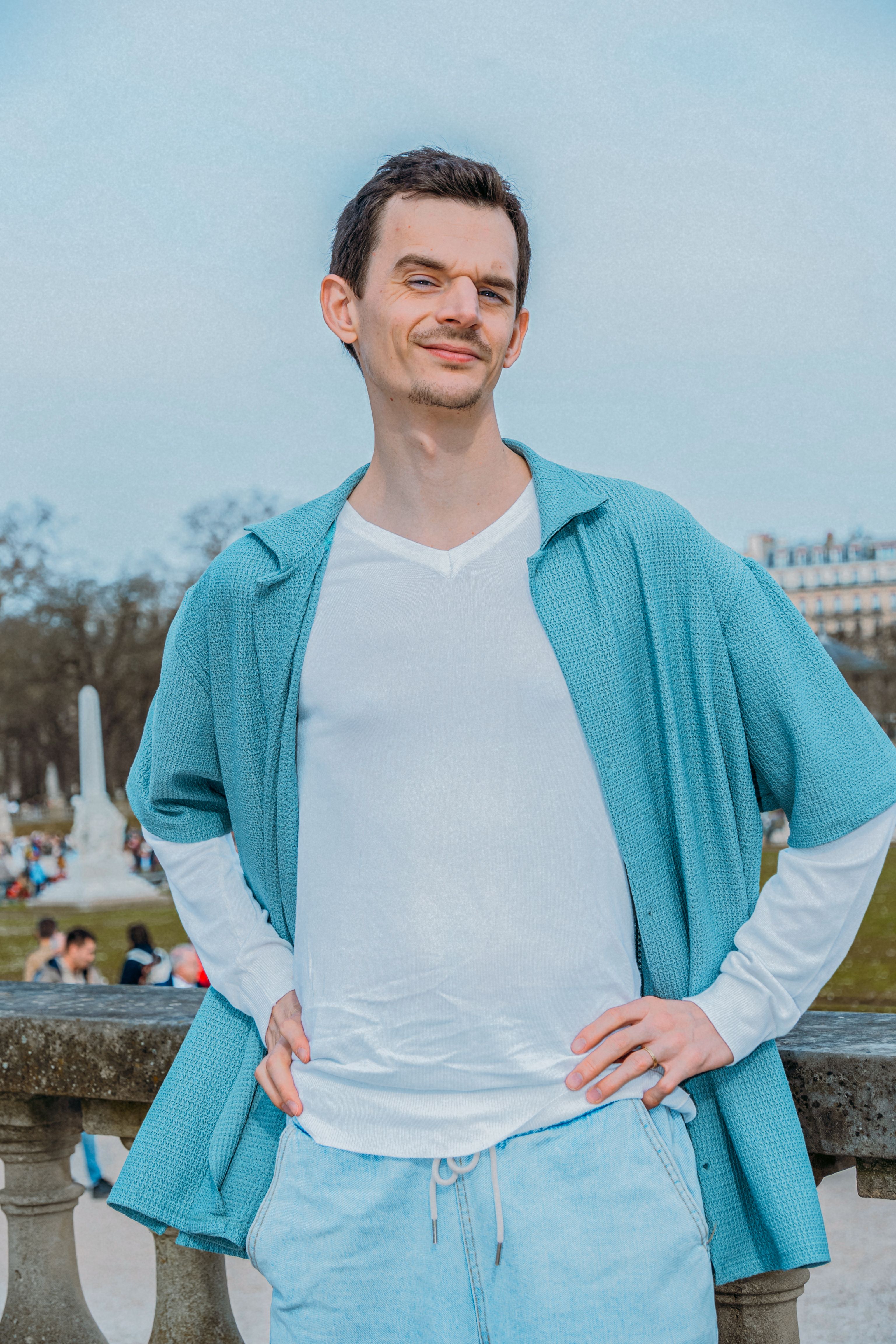 Confident young man poses in casual attire at a park in Paris, showcasing street fashion style.