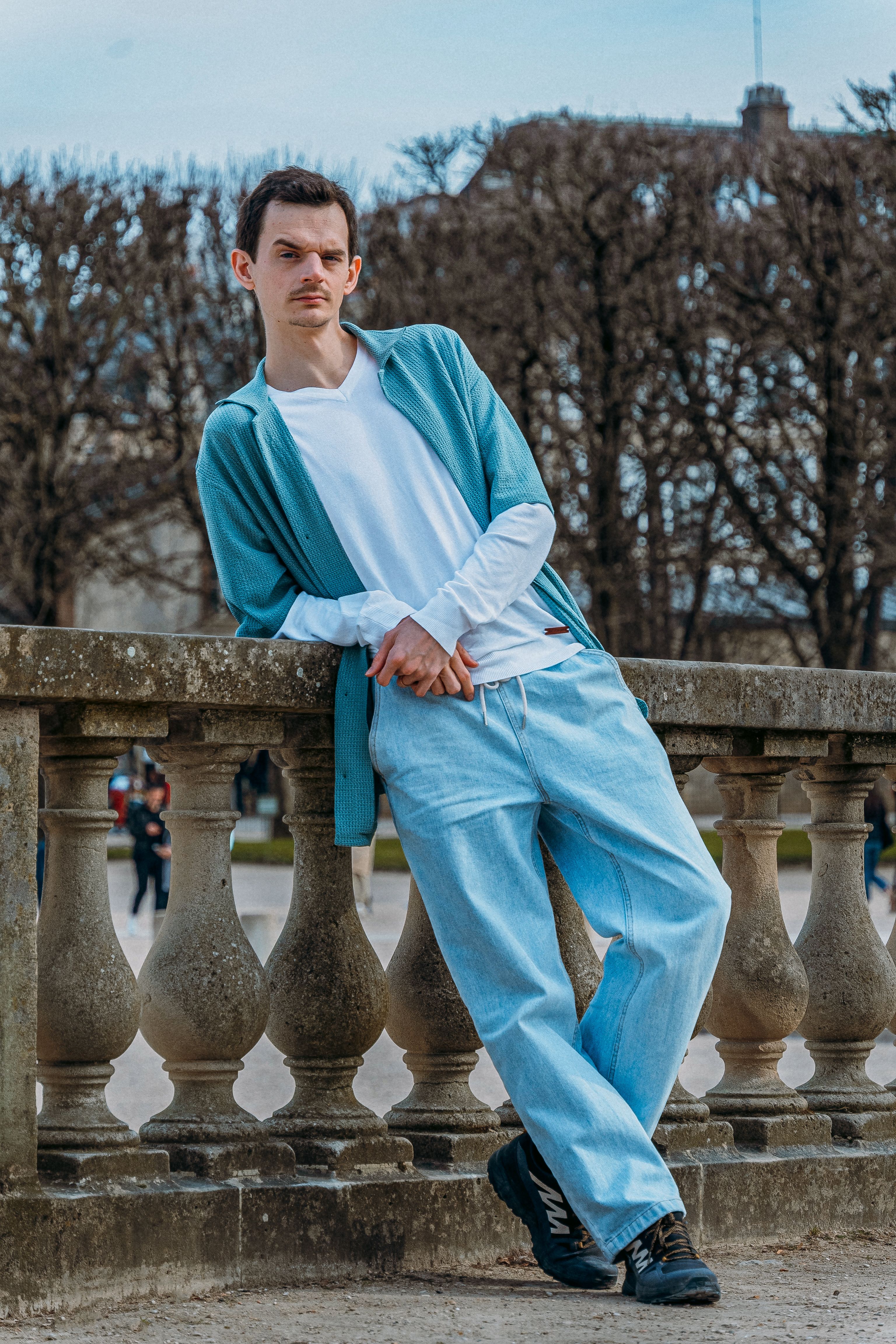A fashionable man in Paris wearing a casual outfit leans against a stone railing.