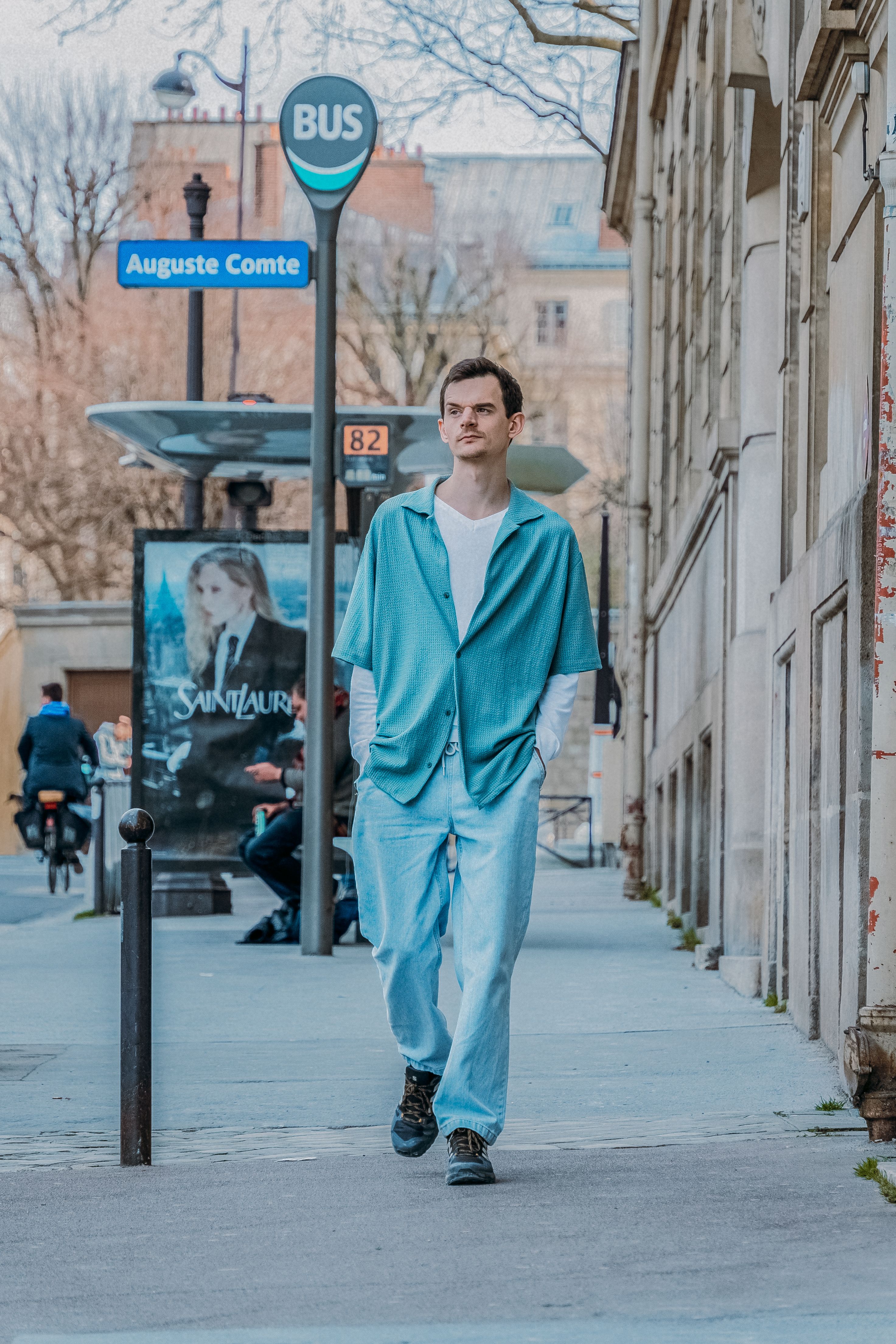 A man showcases street style fashion while wandering through a Parisian street near a bus stop.