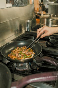 A person stir-frying beef and vegetables in a frying pan on a gas stove.