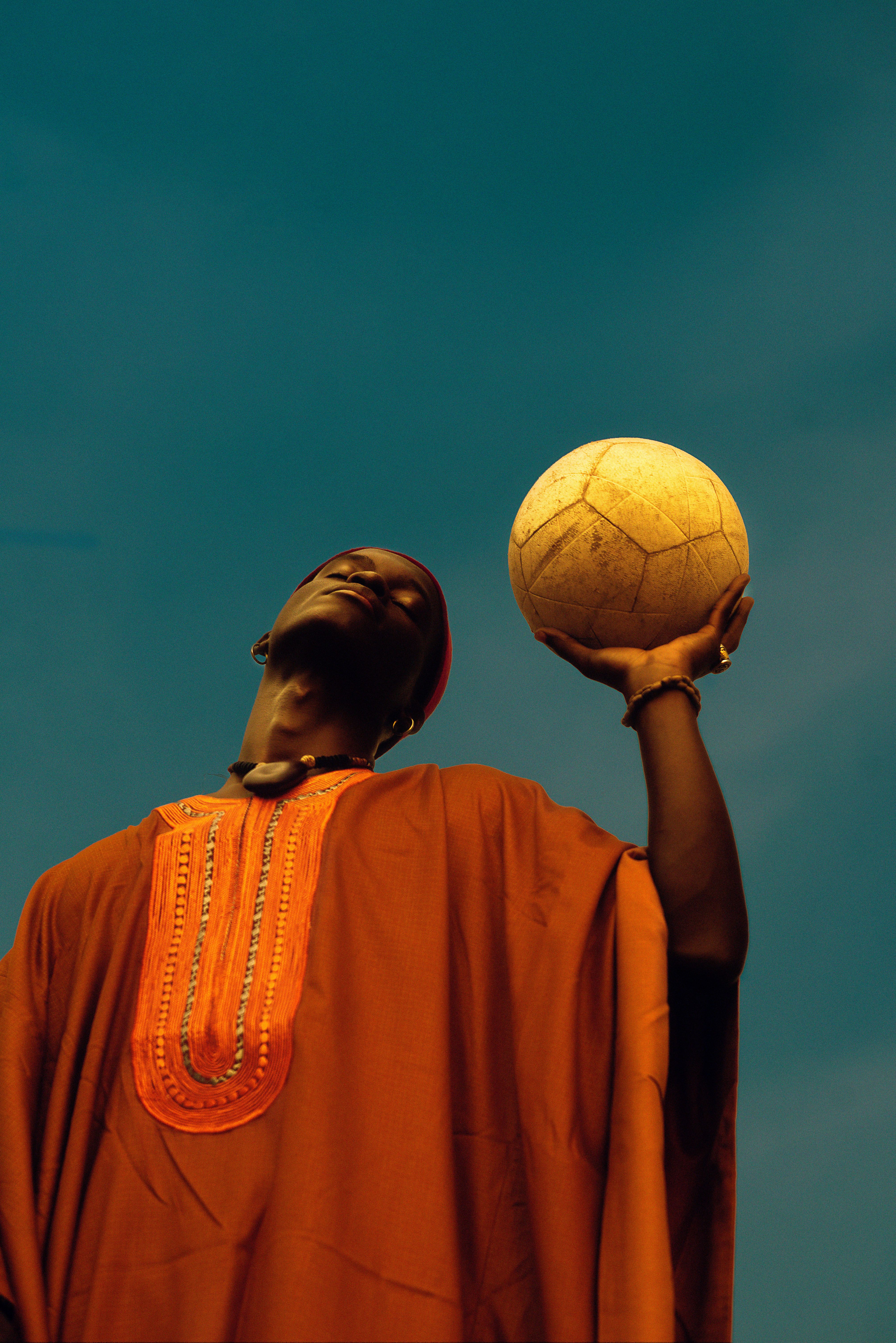 Individual in cultural attire holding a soccer ball against a blue sky.