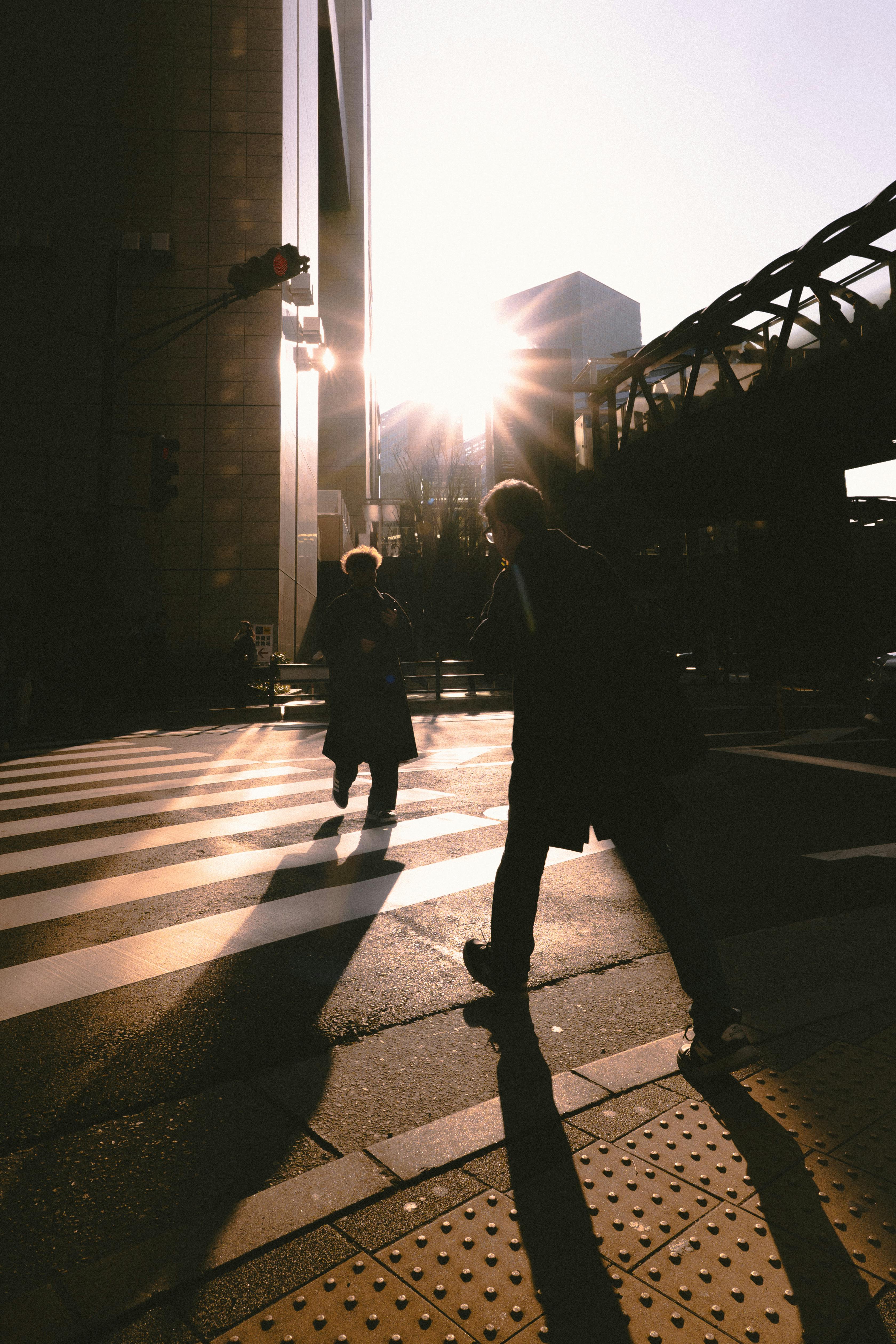 Silhouettes of two pedestrians crossing a city street at sunset with long shadows.