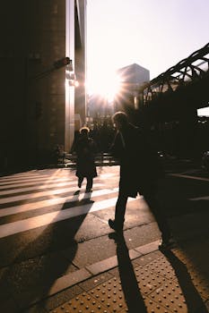 Silhouettes of two pedestrians crossing a city street at sunset with long shadows.