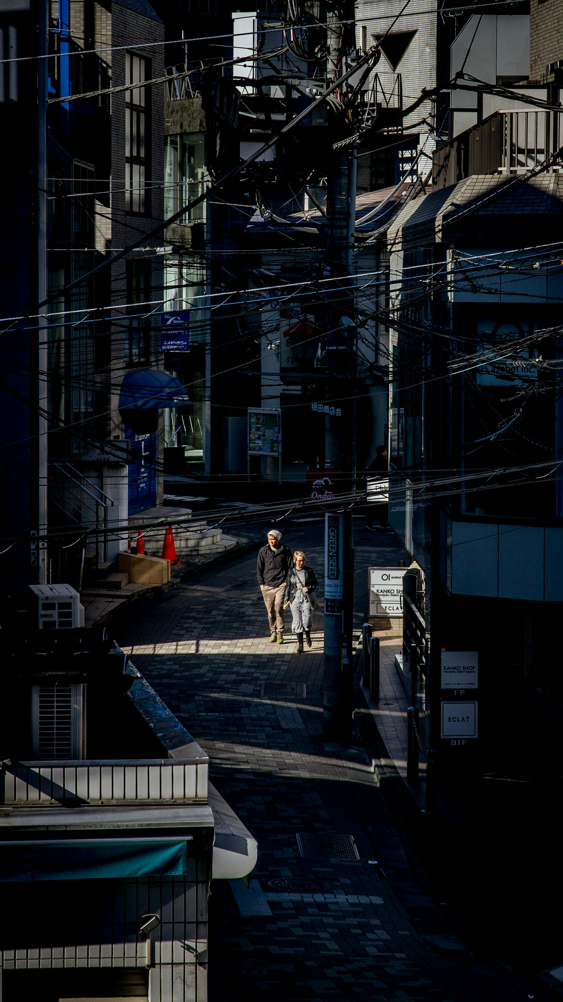 A couple walks down a shadowy urban street amidst complex power lines.