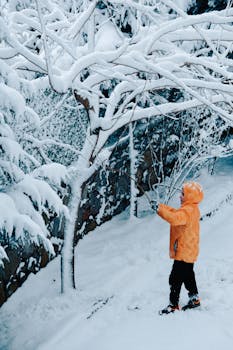 A child in a bright orange jacket plays with snow-covered trees in an İstanbul park during winter.