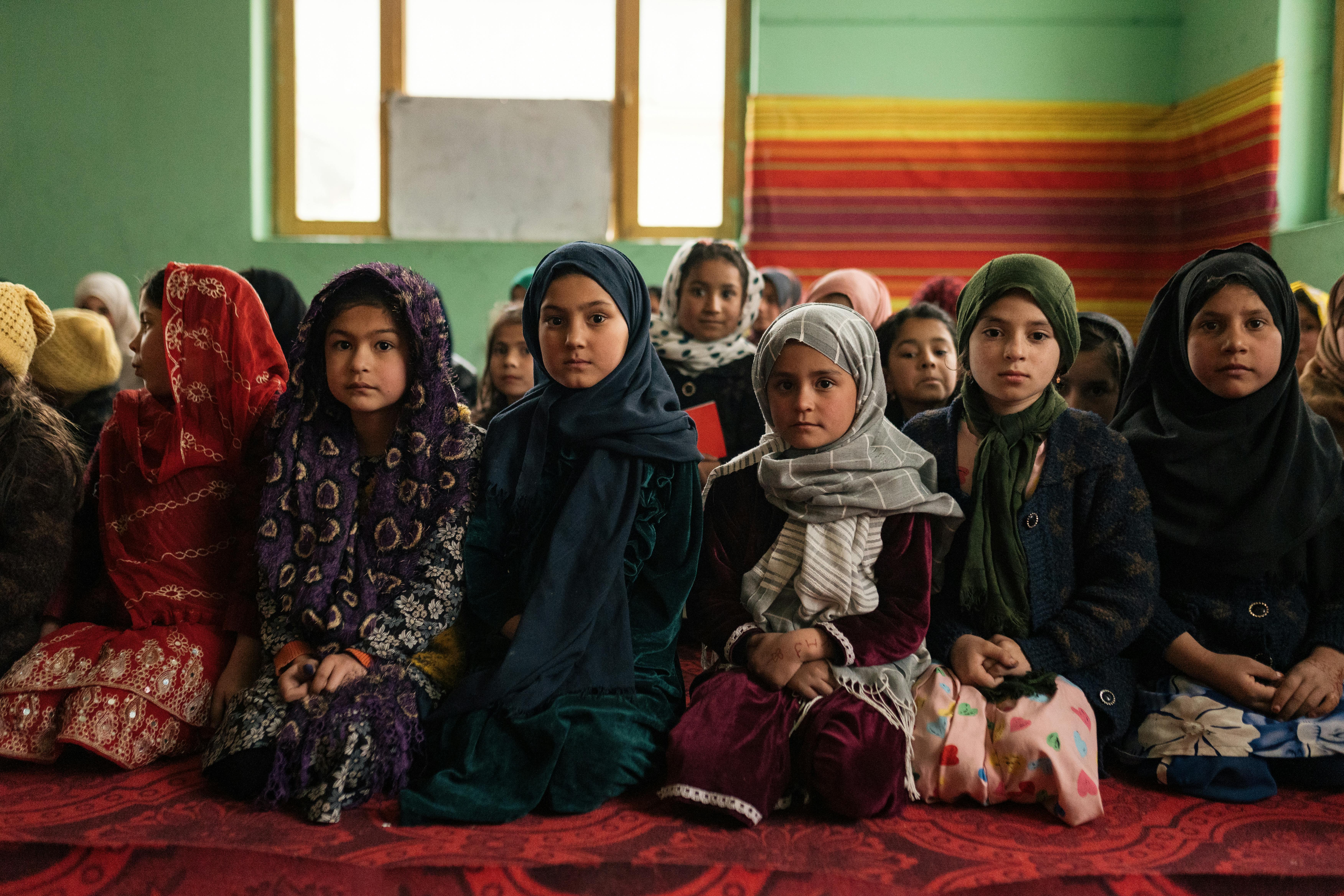 A group of Afghan girls sit indoors, wearing traditional clothing and headscarves.