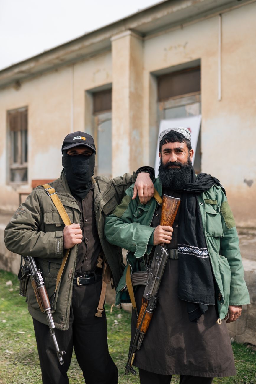 Twee gewapende mannen poseren voor een gebouw en tonen traditionele kledij en wapens uit Afghanistan.