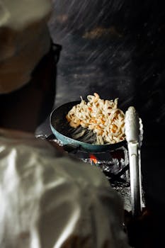 A chef cooks noodles in a dimly lit kitchen setting, showcasing culinary skills.