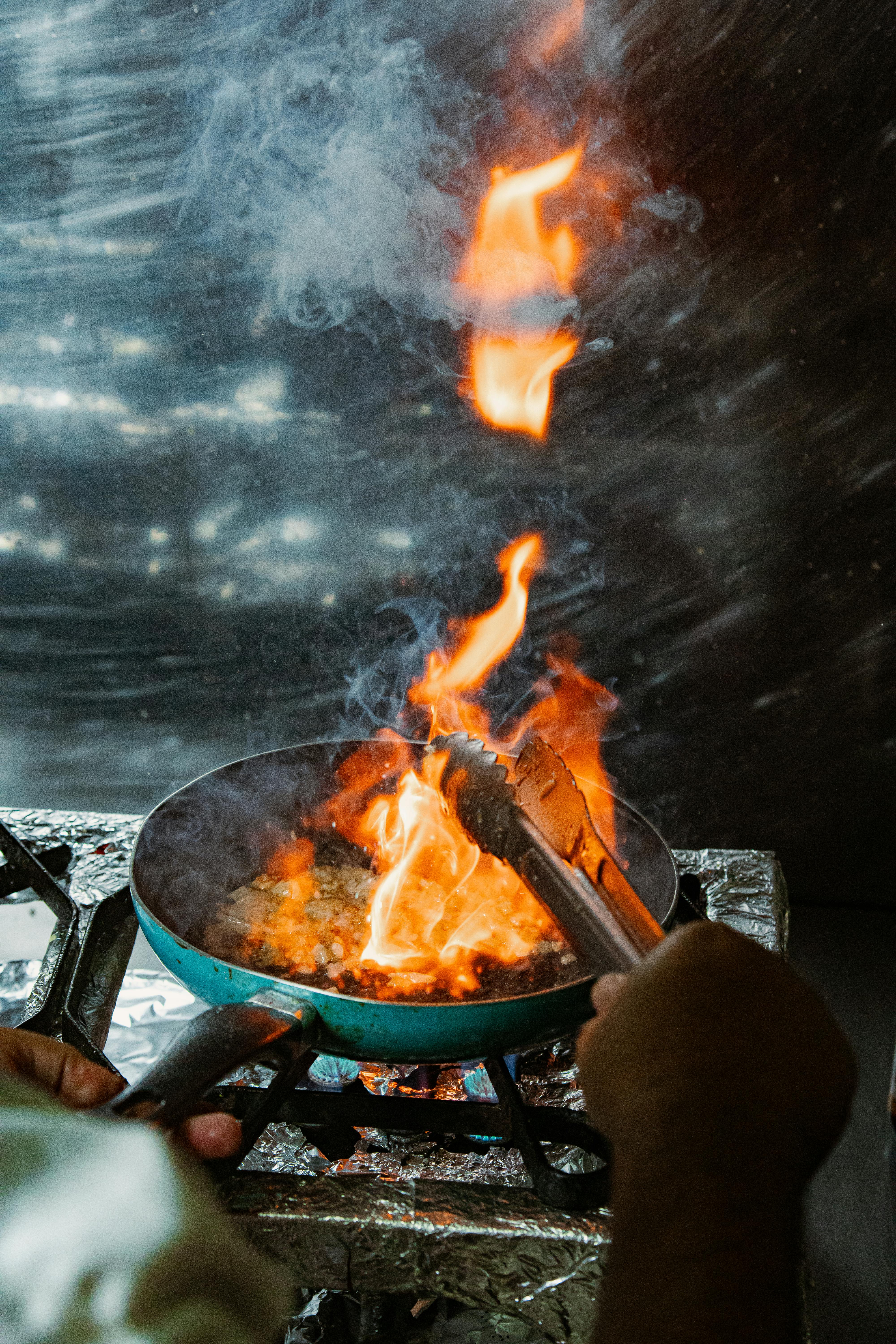 Chef using tongs to flambé dish on stove, dramatic fire for culinary skills.