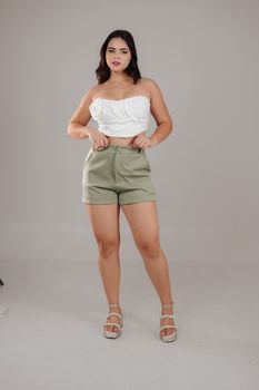 Confident woman in white top and green shorts posing indoors with a neutral background.
