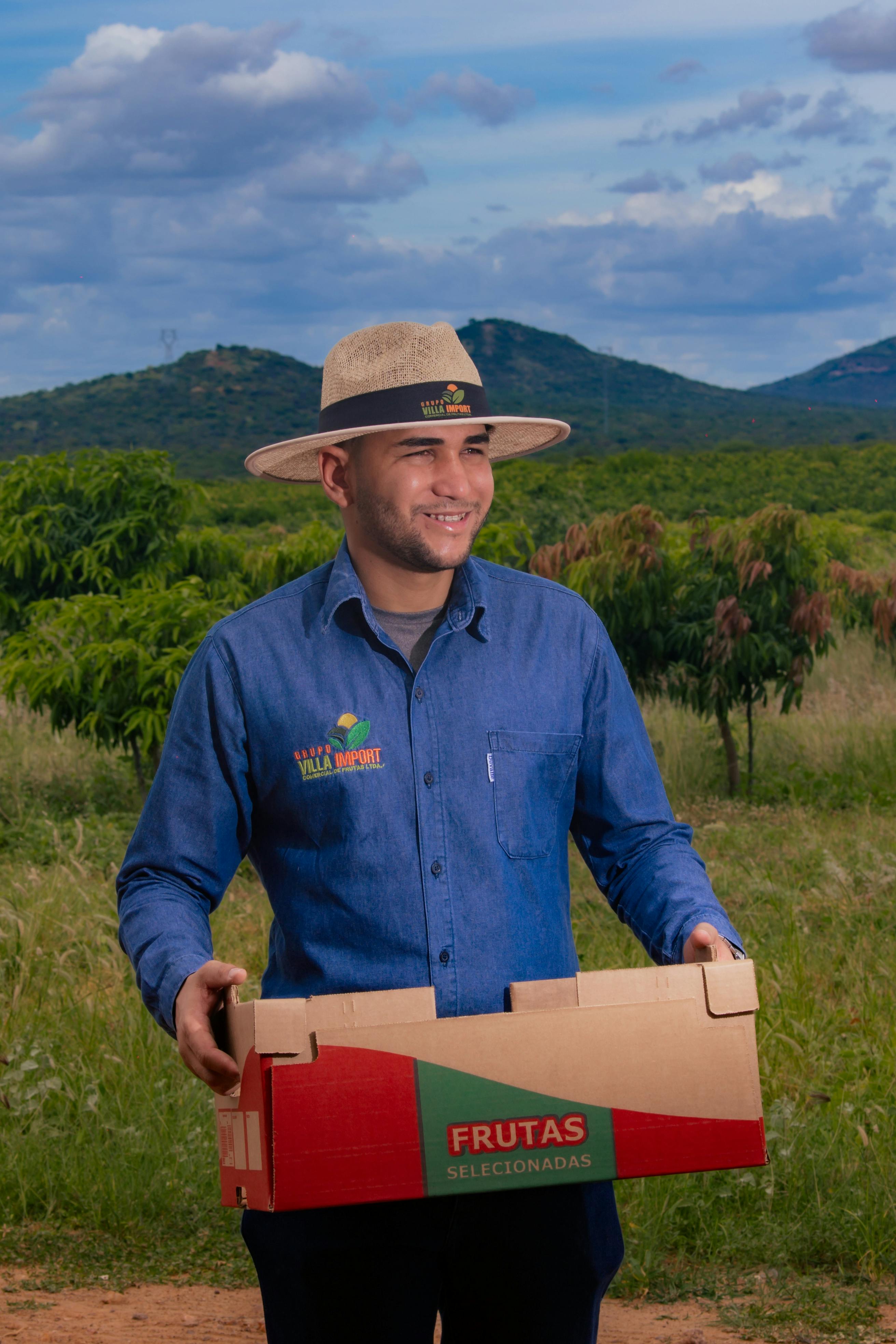 A smiling male farmer in a denim shirt holds a box of fruit outdoors, wearing a straw hat.