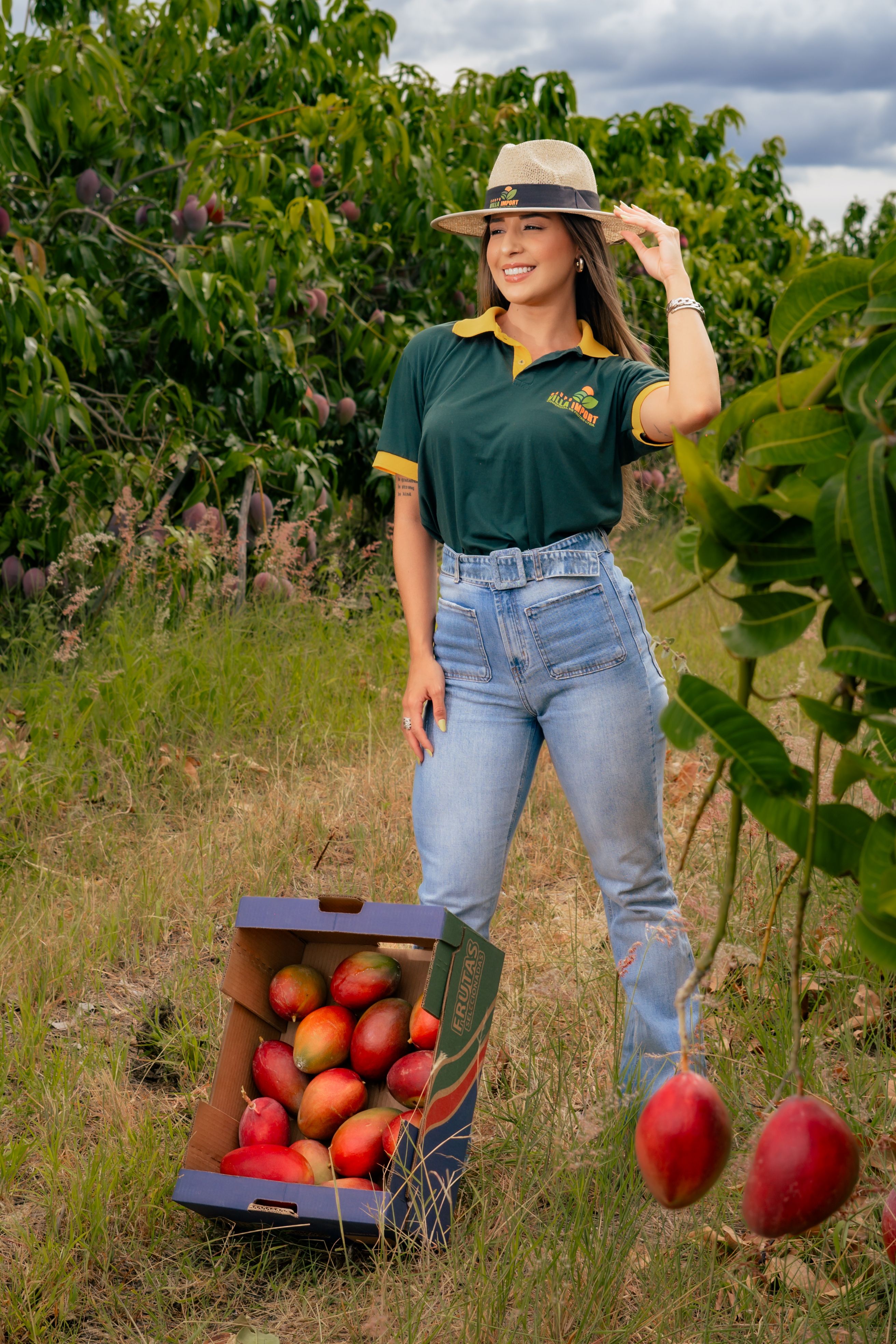 Smiling woman in a mango orchard holding a hat beside a box of ripe mangoes.
