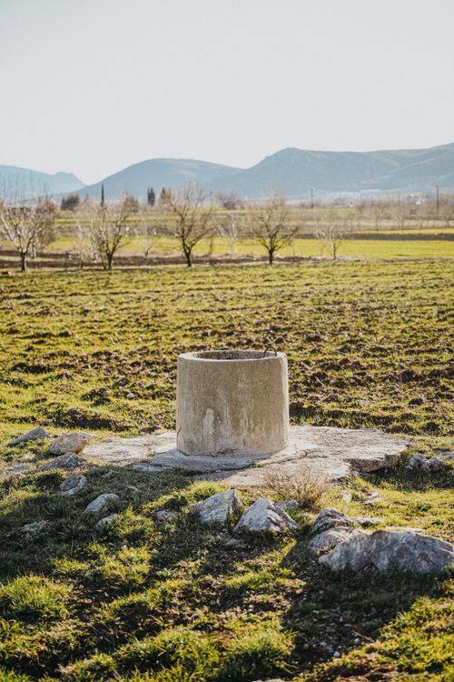 Free Serene rural landscape featuring an old stone well amidst a vast green field and distant mountains. Stock Photo