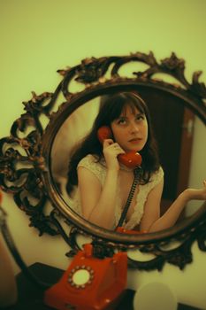 A woman in vintage style using a red rotary phone reflected in an ornate mirror.