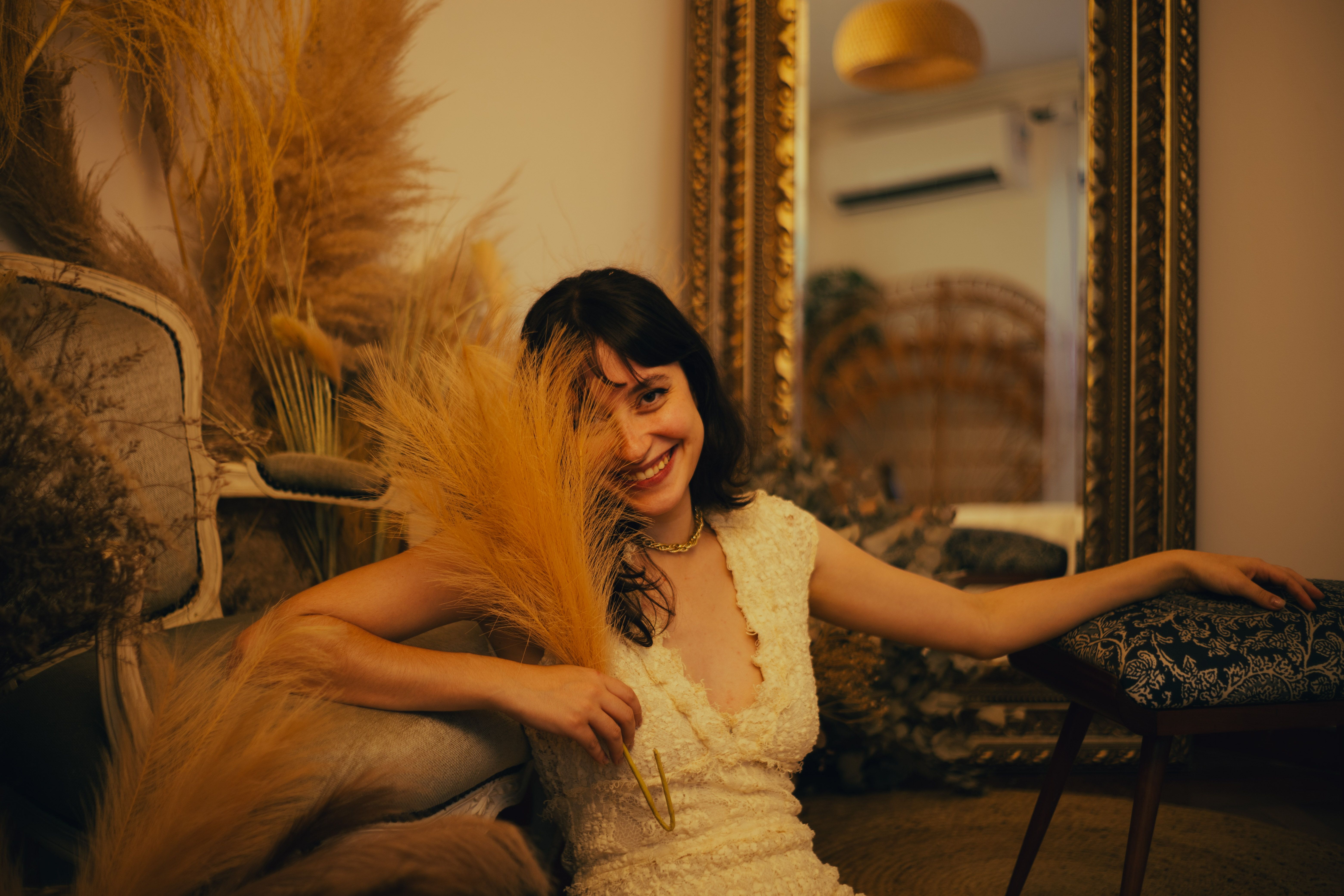 Smiling woman in white dress surrounded by pampas grass indoors.